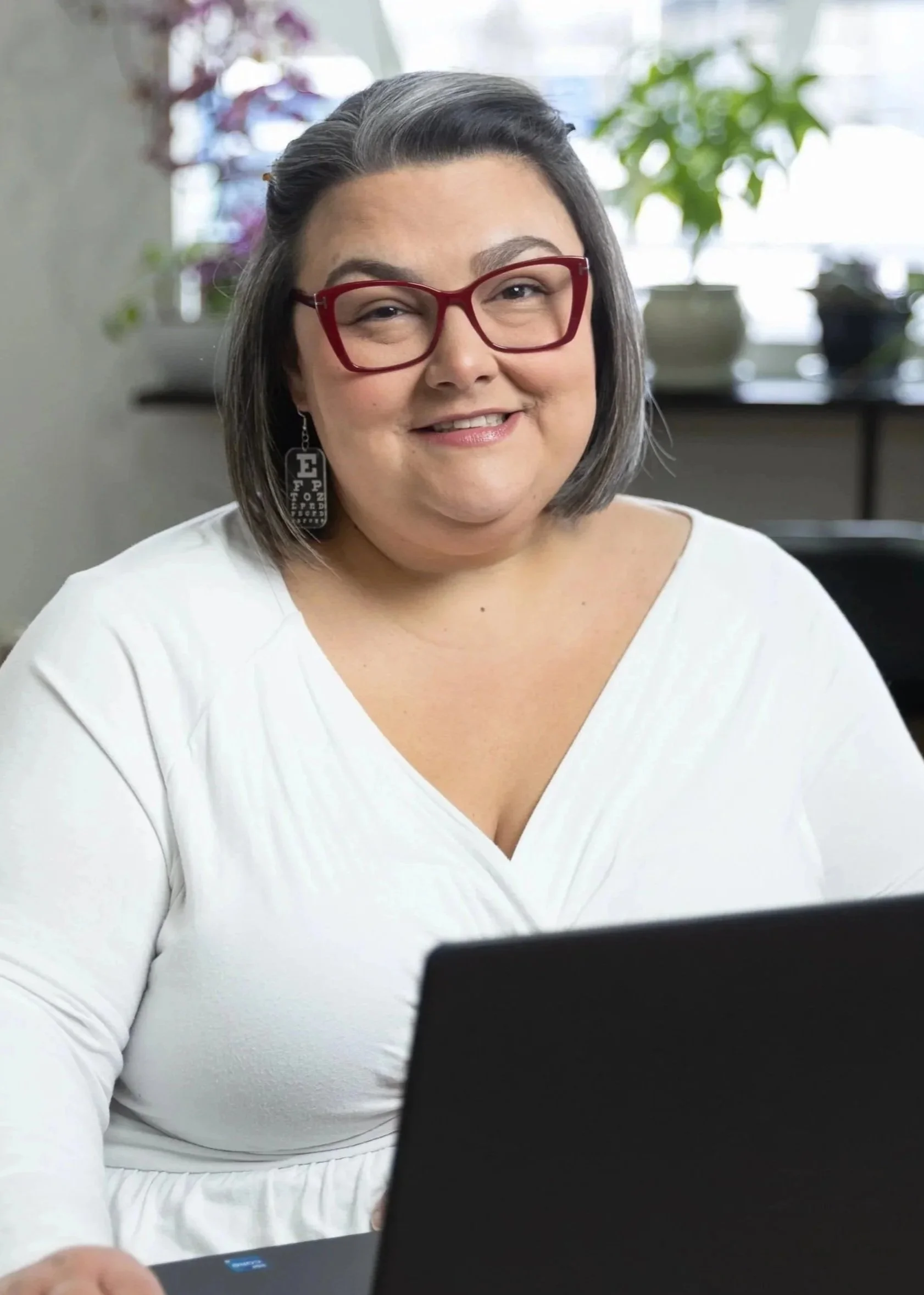 A woman wearing red glasses and earrings smiling while working on a laptop in a room with plants and natural light.