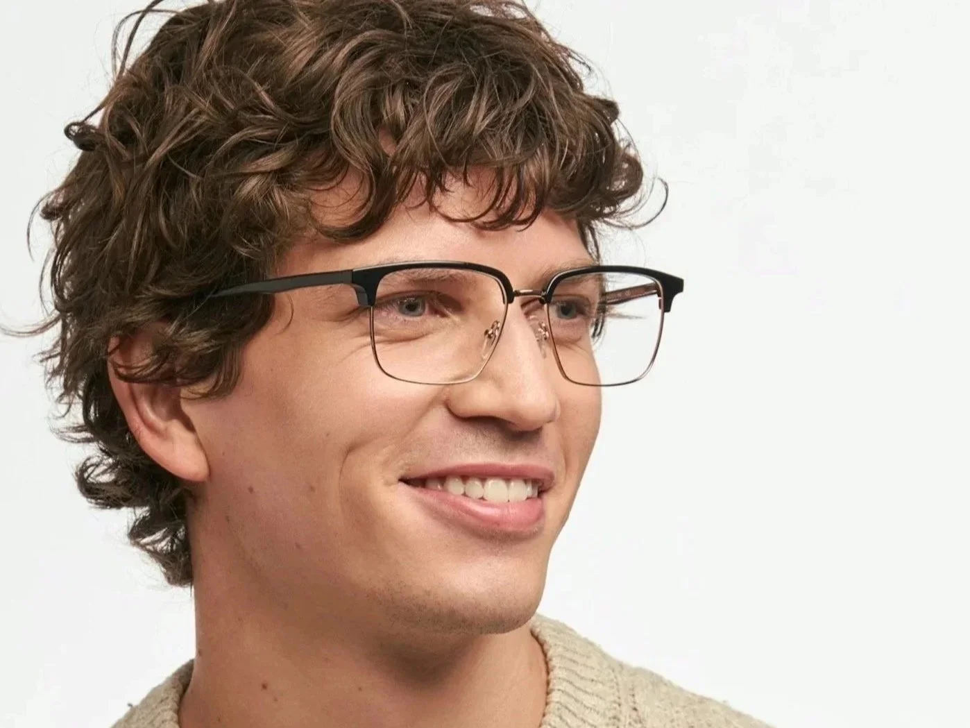 Close-up of a young man with brown curly hair, wearing black-rimmed glasses and a light-colored sweater, smiling against a plain background.