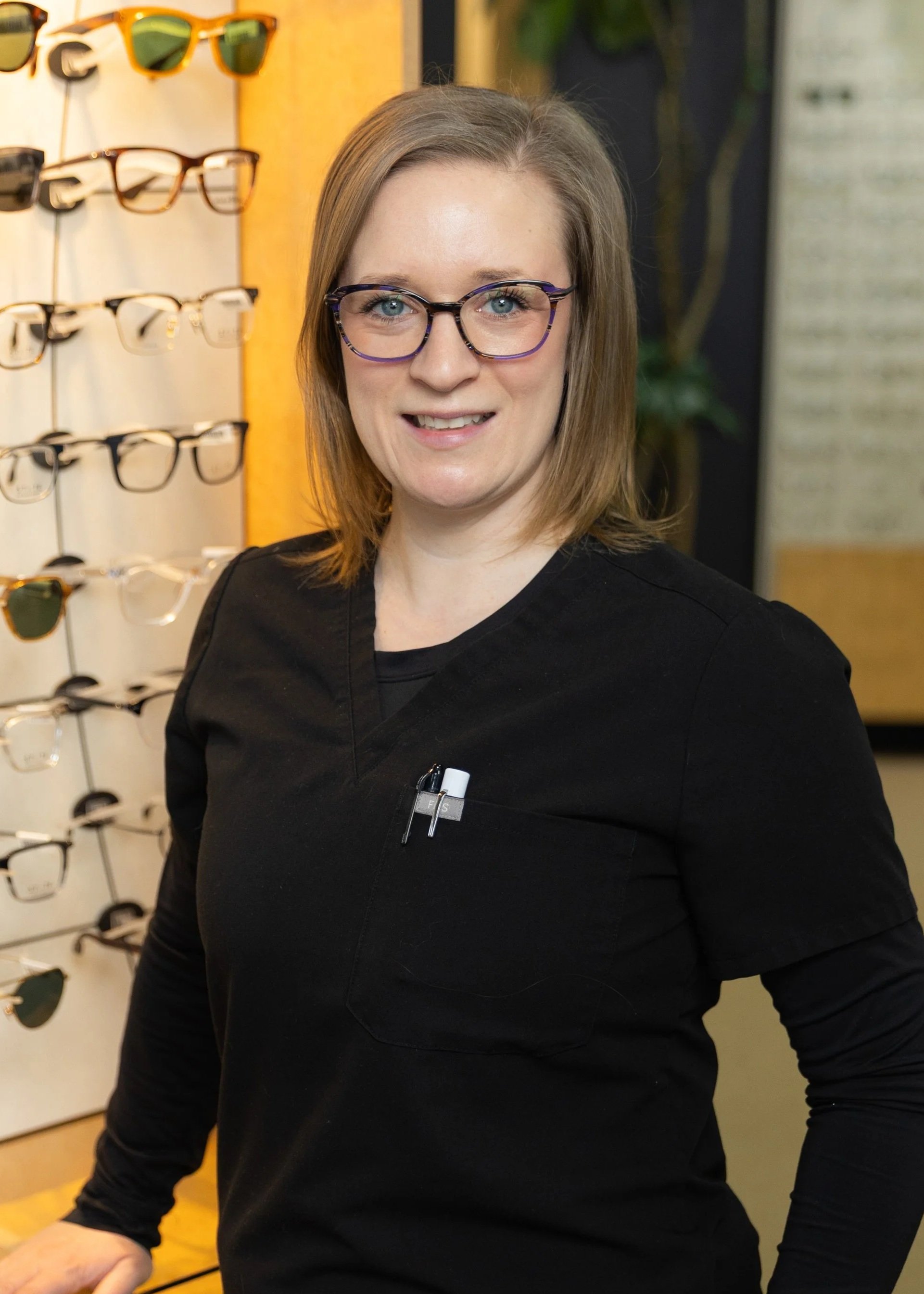 A woman with shoulder-length blonde hair wearing glasses and a black work uniform with pens and a white marker in the chest pocket. She is standing in an eyewear store with a display of glasses behind her.