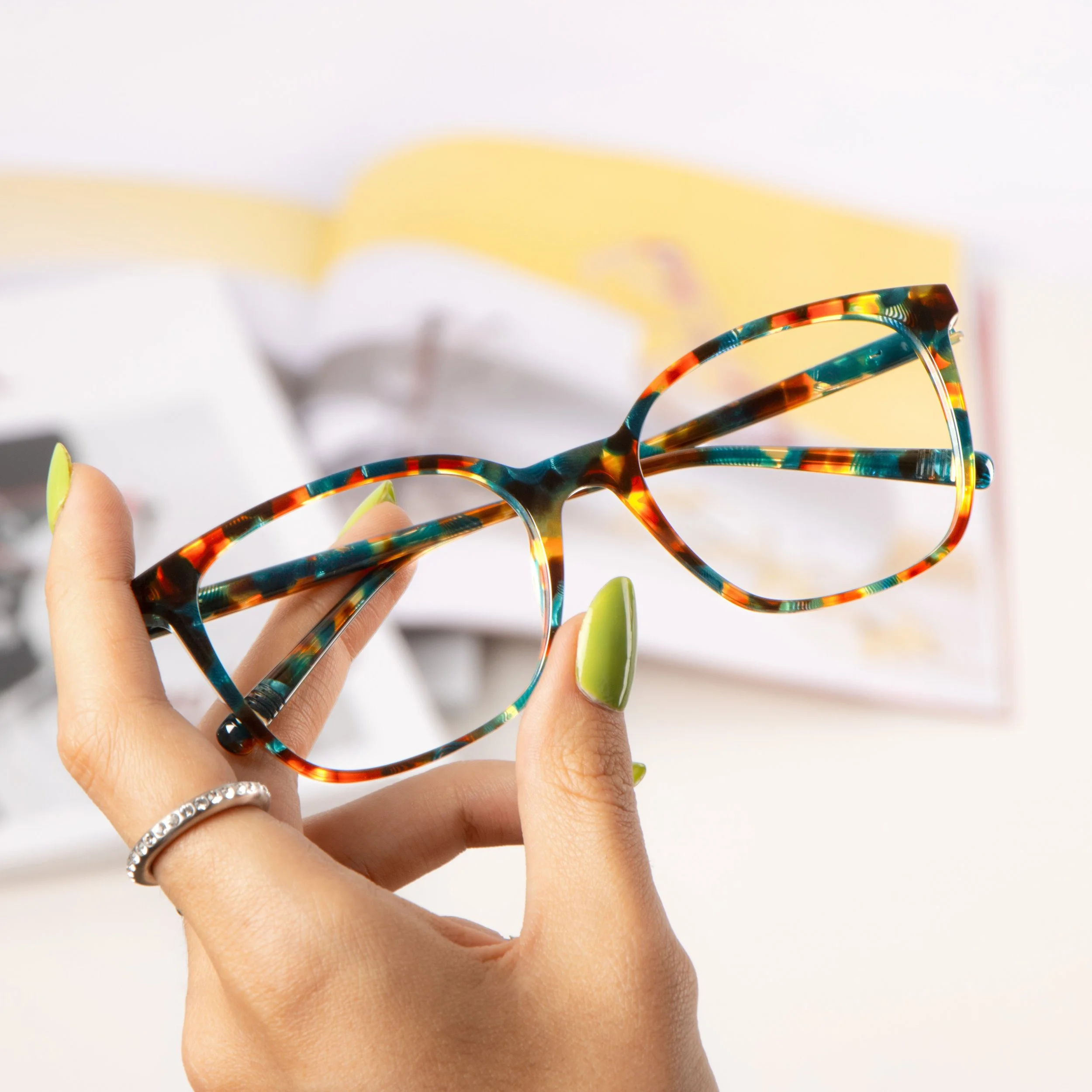 Person holding colorful tortoiseshell glasses against a light background, with nails painted light green and rings on fingers.