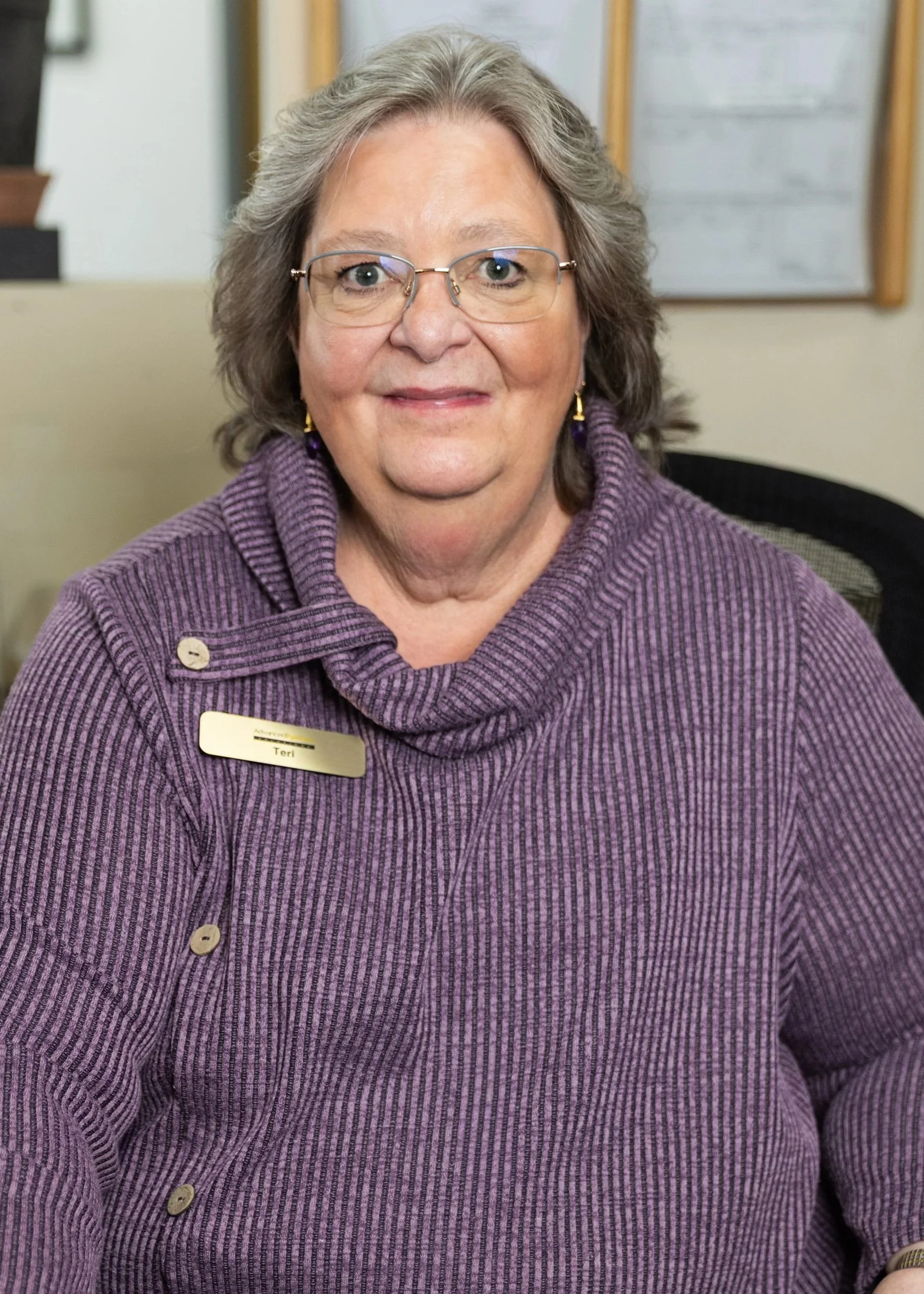 A middle-aged woman with gray hair and glasses, wearing a purple knitted top with a name tag that reads 'Teri', sitting indoors in front of a bulletin board.