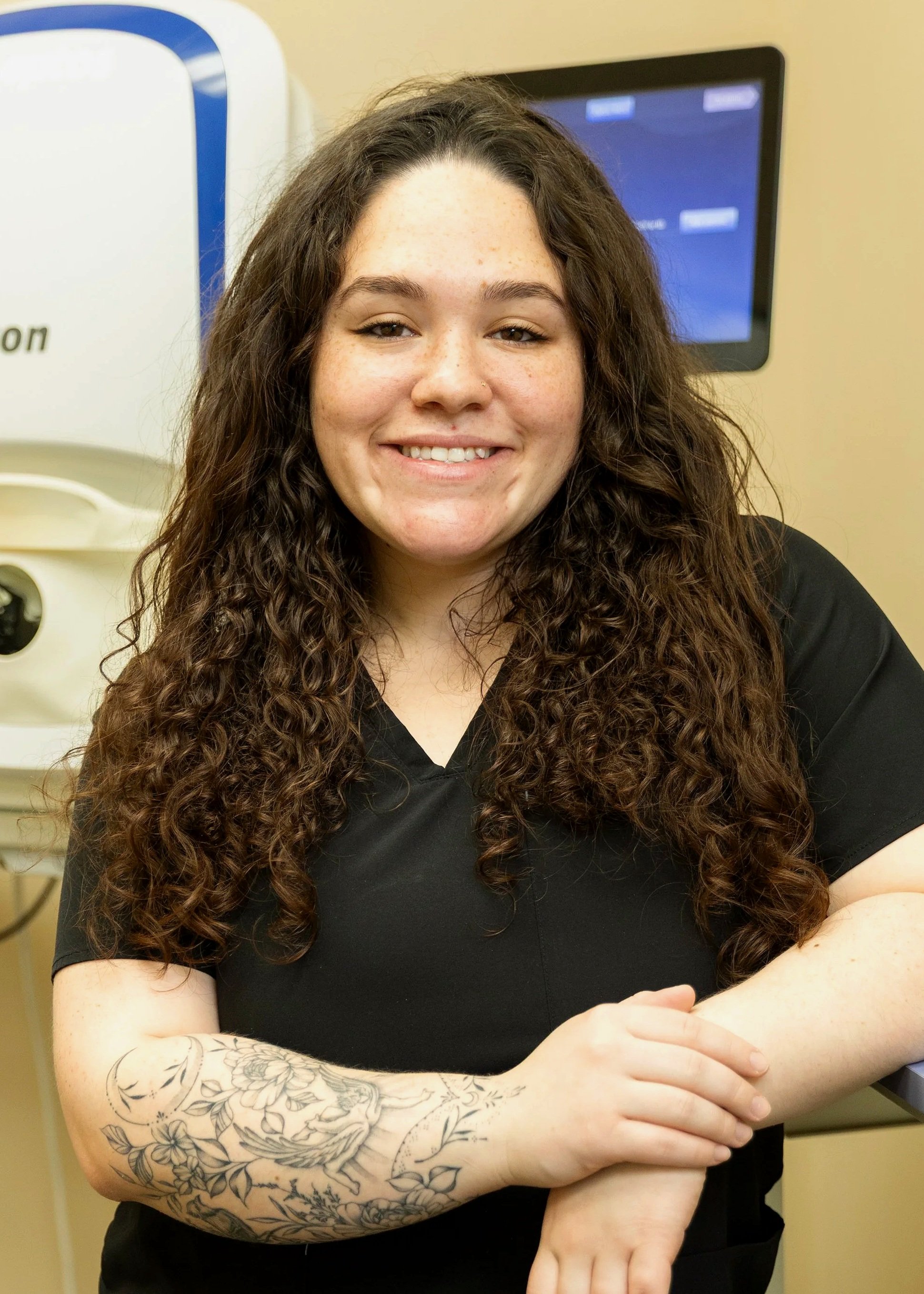 A woman with long, curly brown hair and a tattoo on her left arm, smiling while sitting in a medical setting with medical equipment and a computer screen in the background.