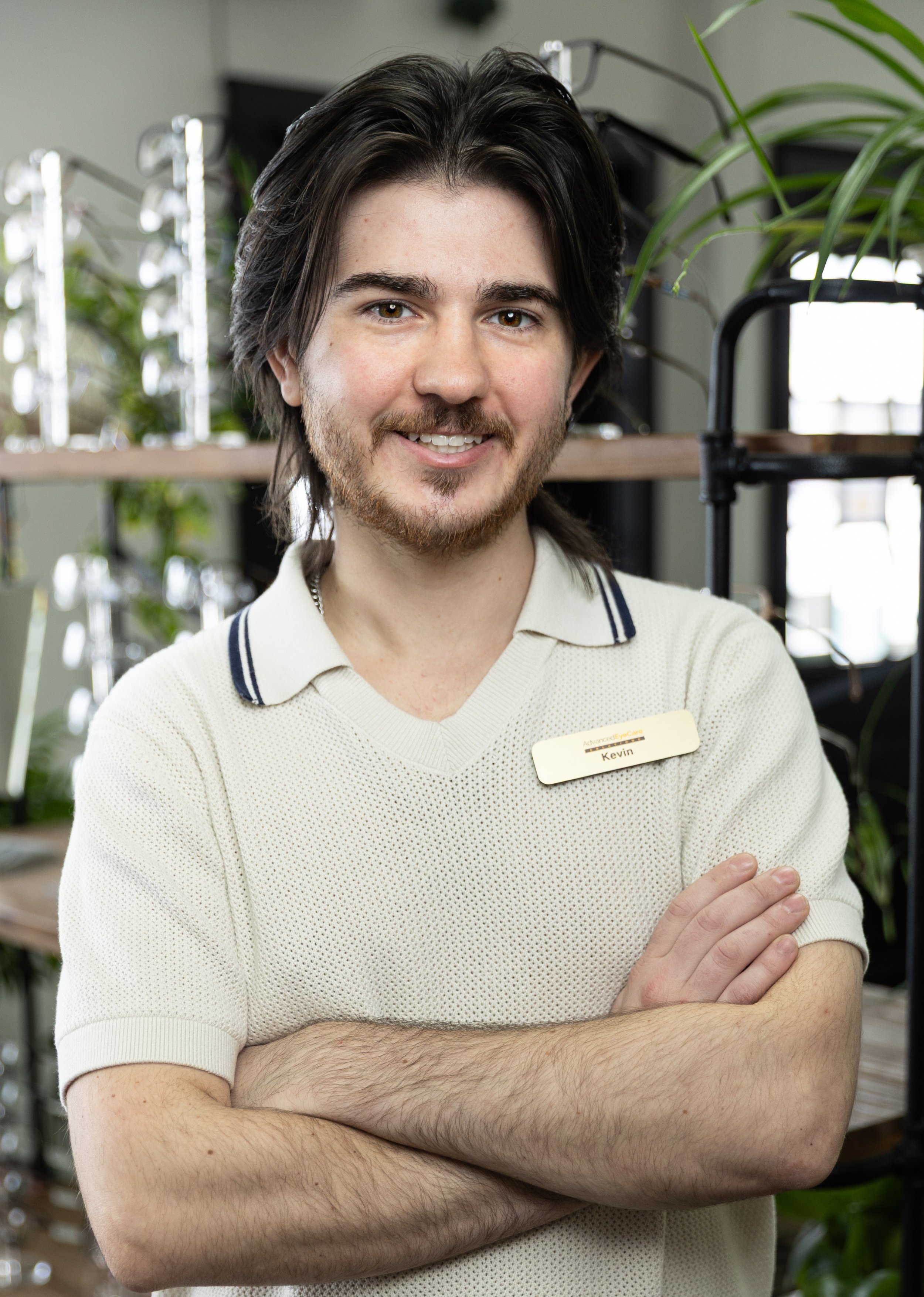 Young man with shoulder-length dark hair and a beard, wearing a cream-colored polo shirt with dark stripes on the collar and a name tag that reads 'Kevin', standing with arms crossed in an indoor setting with plants and shelves in the background.