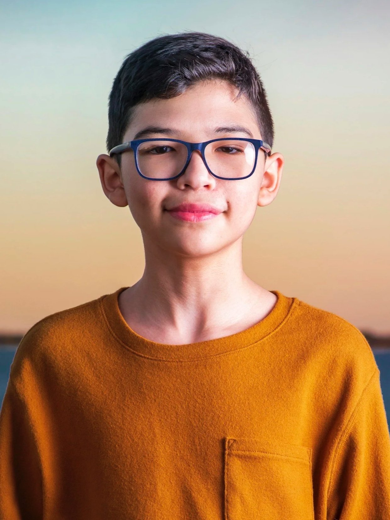 A young boy with short dark hair, glasses, wearing an orange sweater, smiling softly against a neutral background.