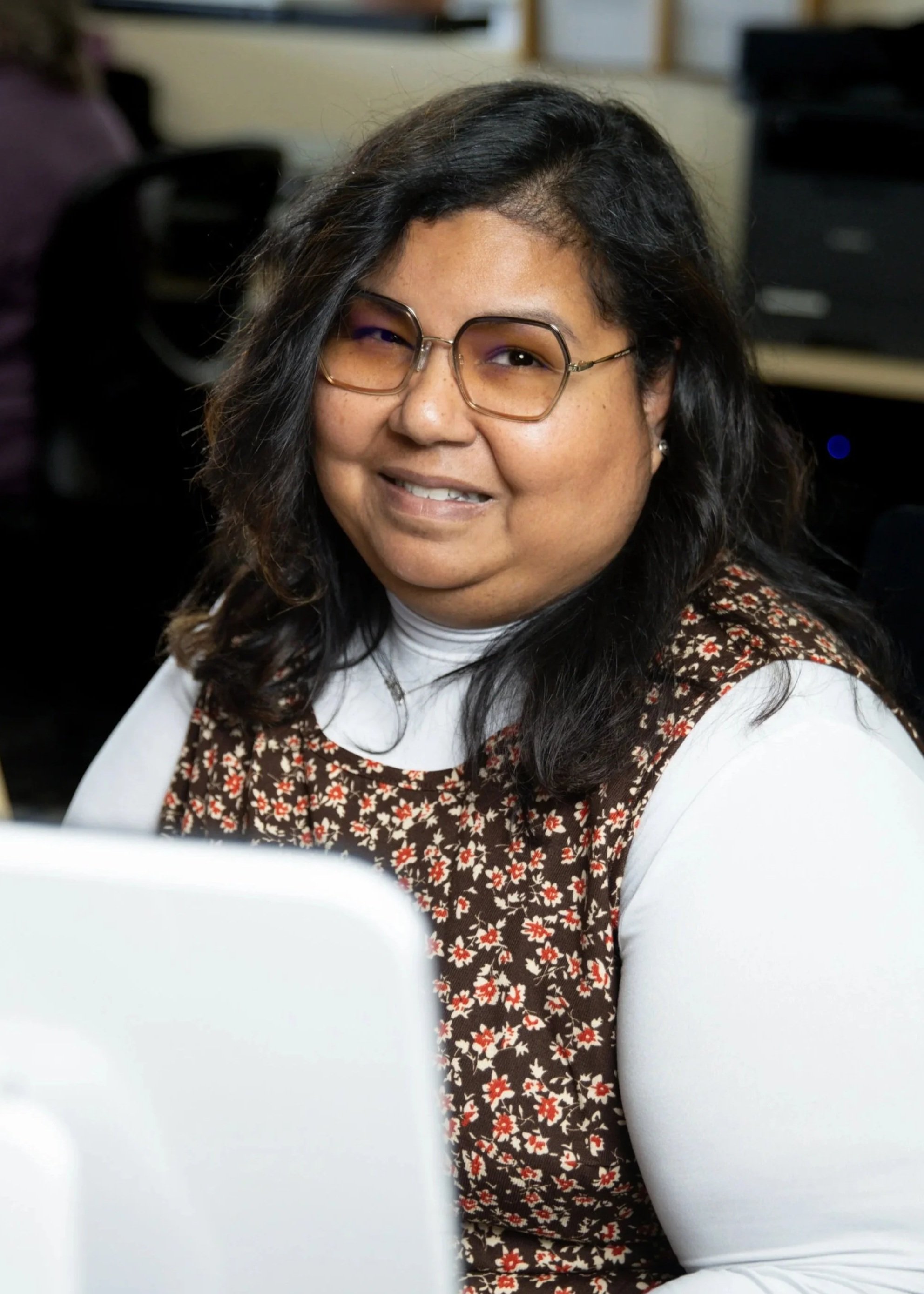 A woman with dark hair, glasses, and a floral brown and white shirt, smiling at a desk in an office setting.