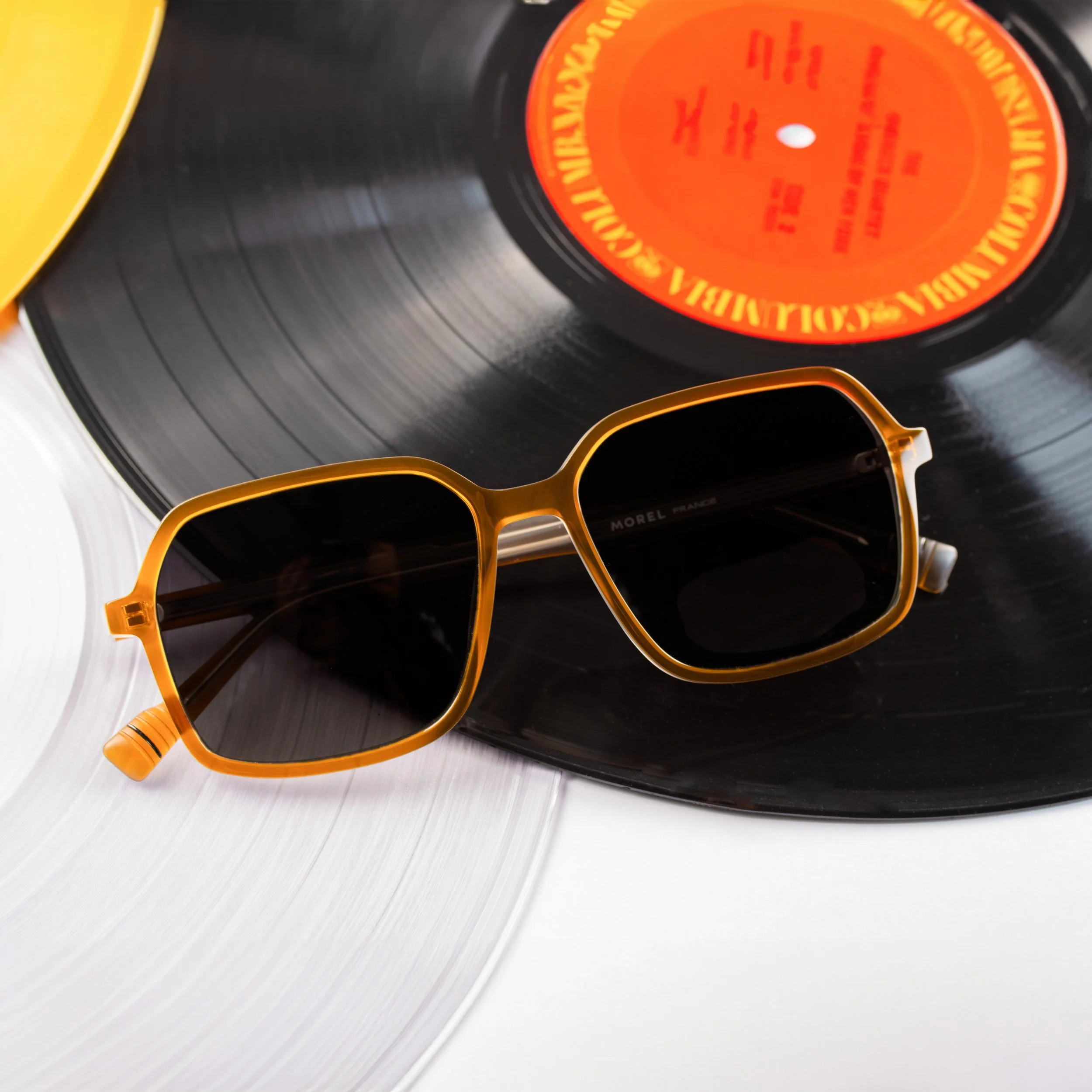 Black vinyl record with orange label and yellow sunglasses resting on it, on a white surface.