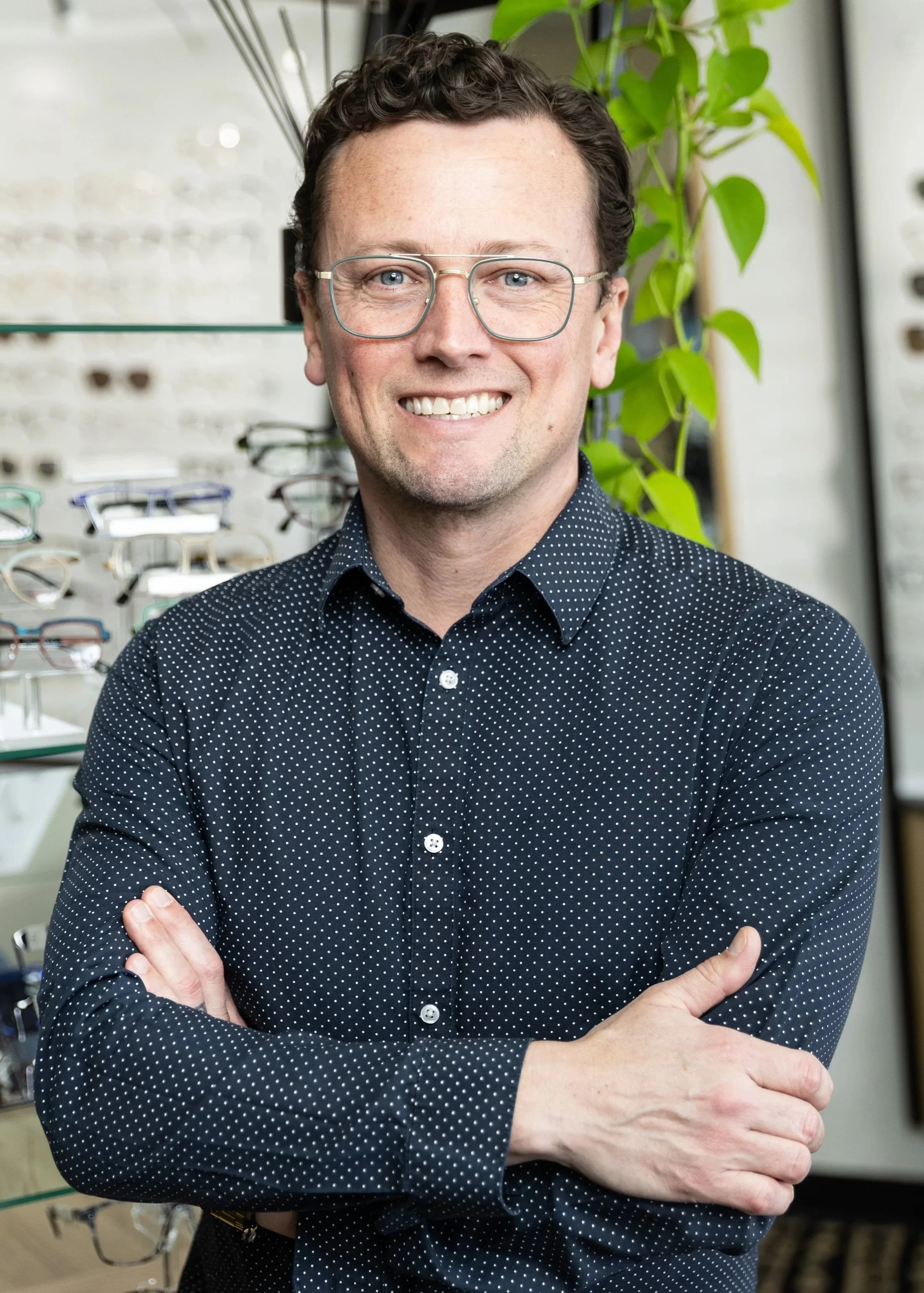 A professional man with brown hair, glasses, and a friendly smile standing indoors with arms crossed. He is wearing a navy blue shirt with small white polka dots. There are eyeglass frames on display behind him and green plants nearby.