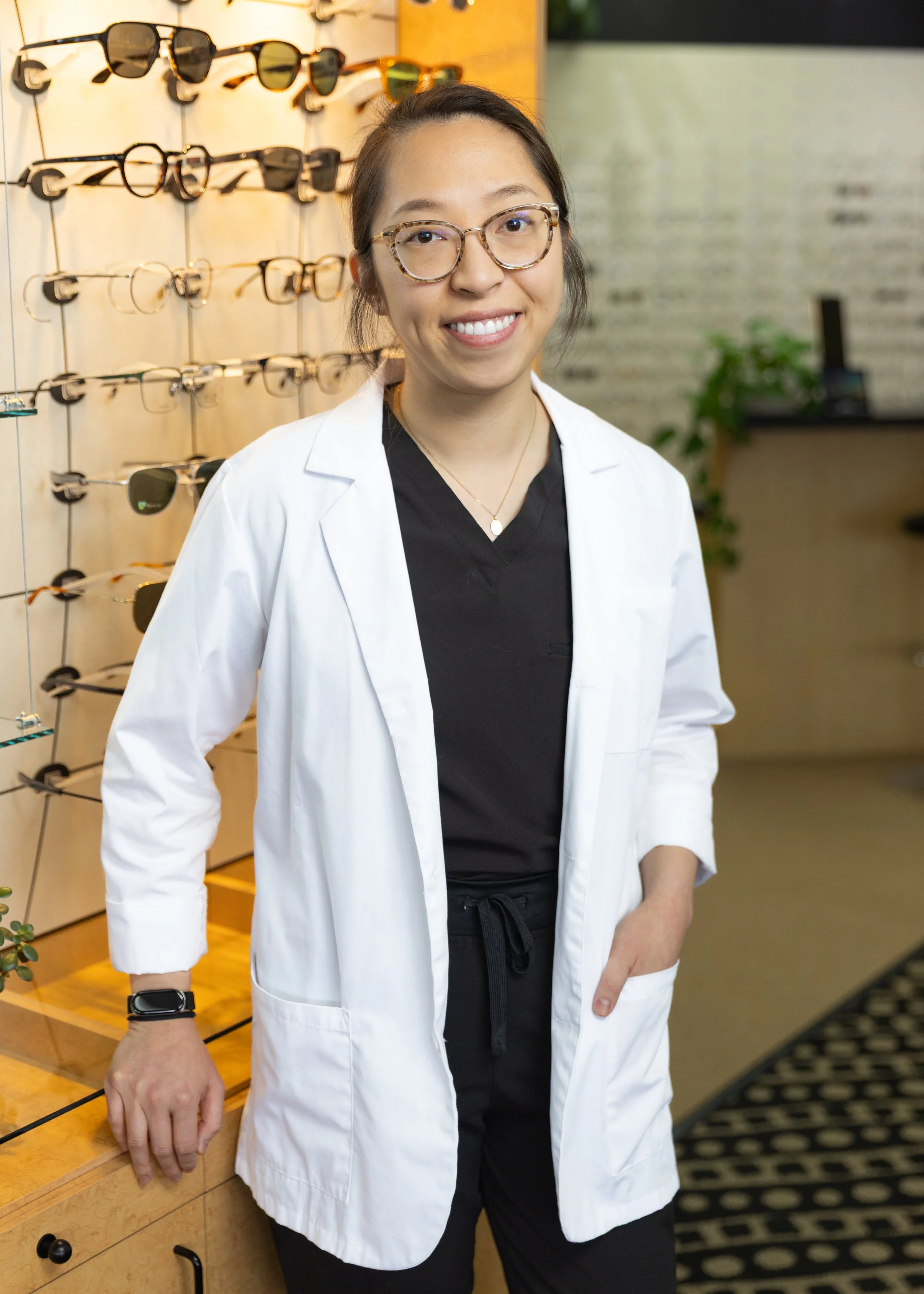 A woman wearing glasses and a white coat smiling inside a store with sunglasses displayed on the wall behind her.