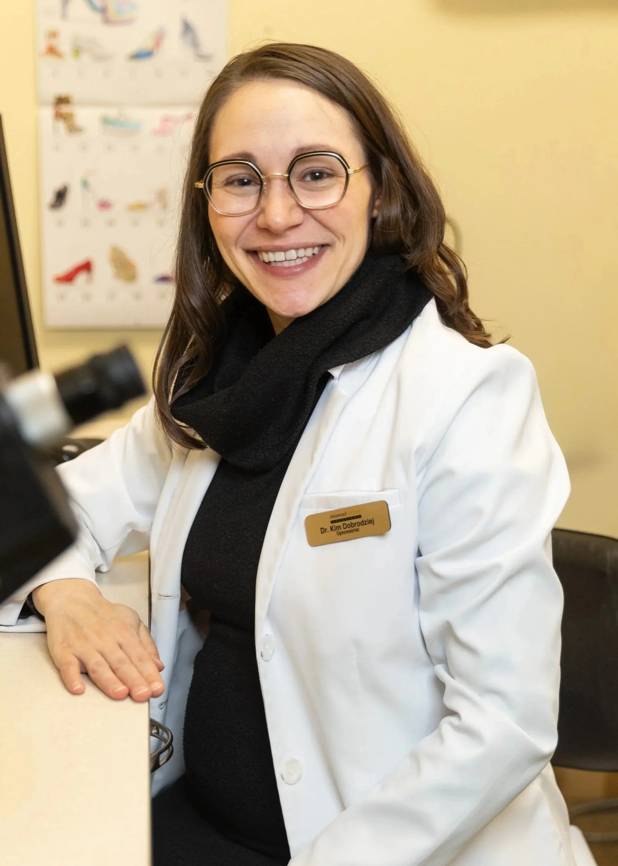 A woman with glasses and brown hair, wearing a white coat and a black scarf, smiling at the camera.