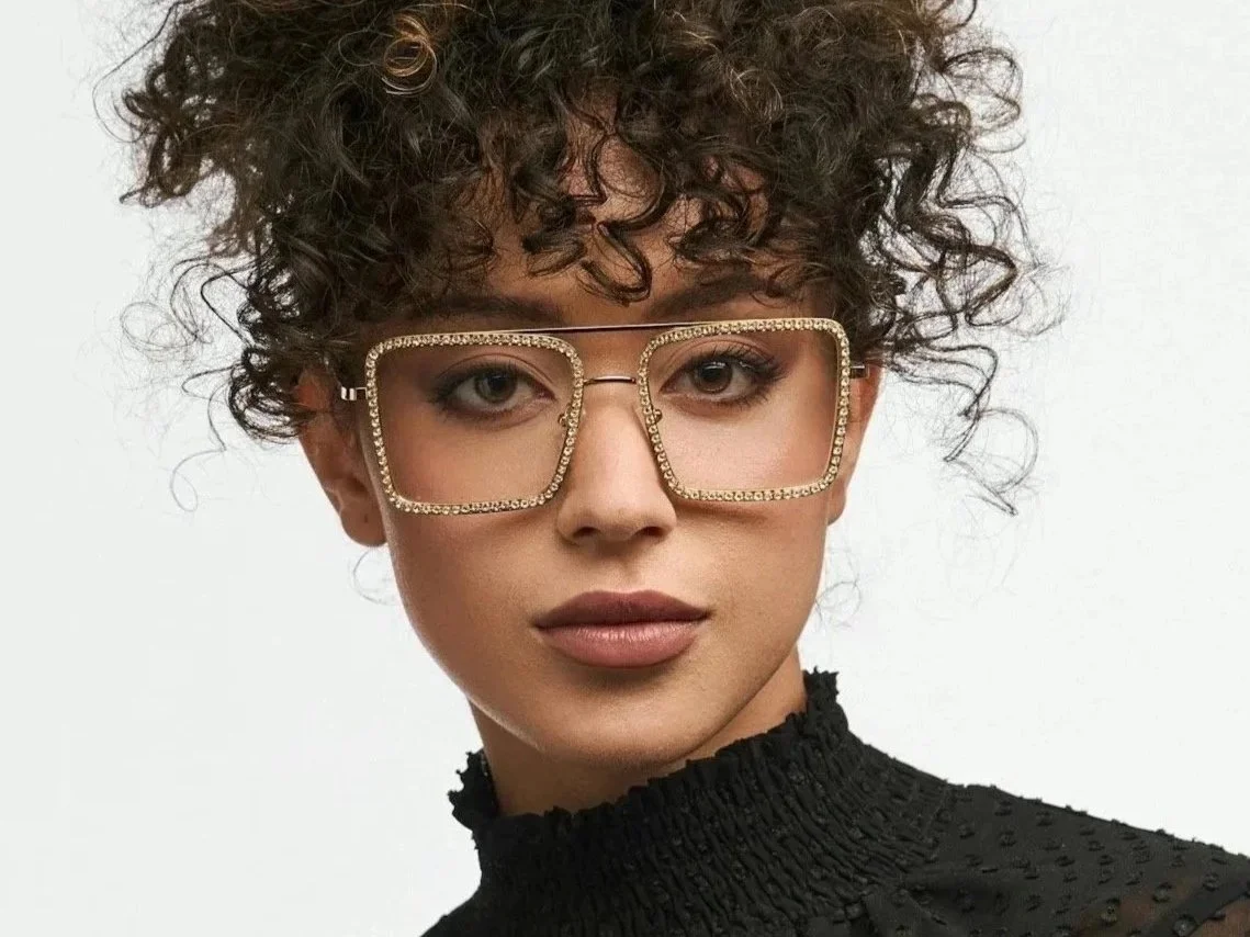 Woman with short, curly hair wearing large, square, rimmed glasses with rhinestones and a black, high-neck textured top.