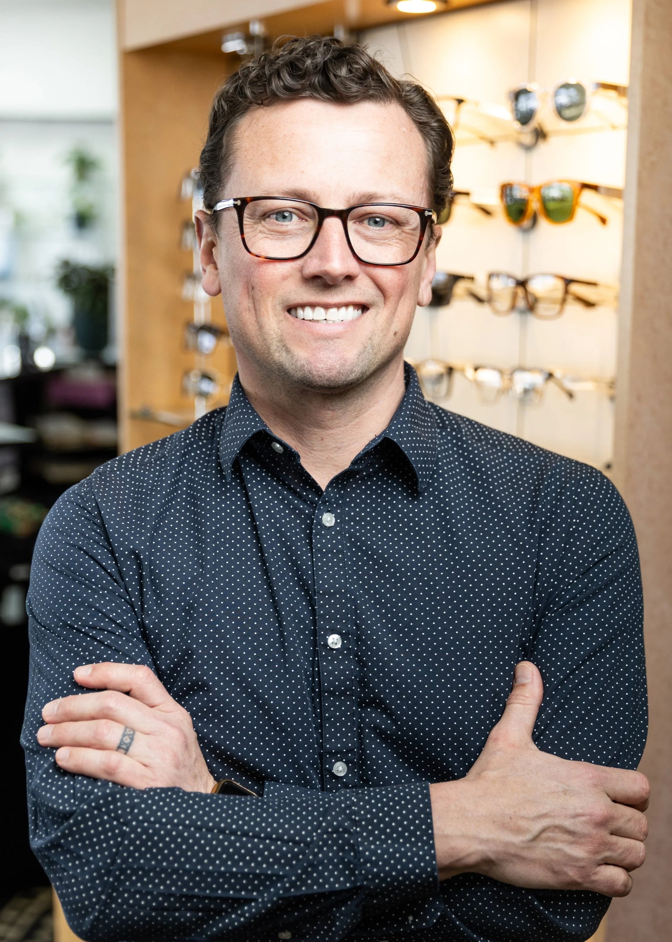 A smiling man with curly hair, glasses, and a dark polka-dot shirt standing indoors with sunglasses display in the background.