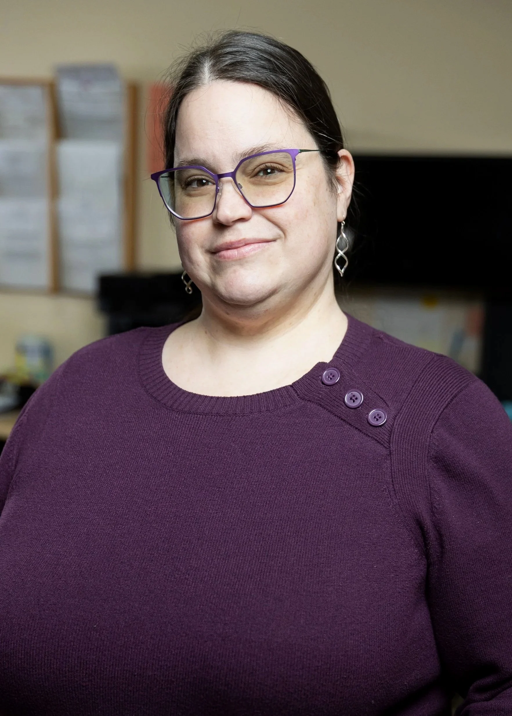 A woman with dark hair, glasses, and earrings, wearing a purple sweater with button details on the shoulder, smiling indoors.