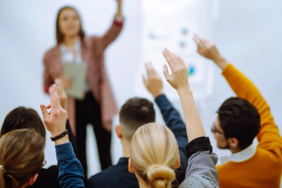 Une femme donne une présentation Qualiopi devant un groupe de personnes, plusieurs participants lèvent la main pour poser une question ou répondre.
