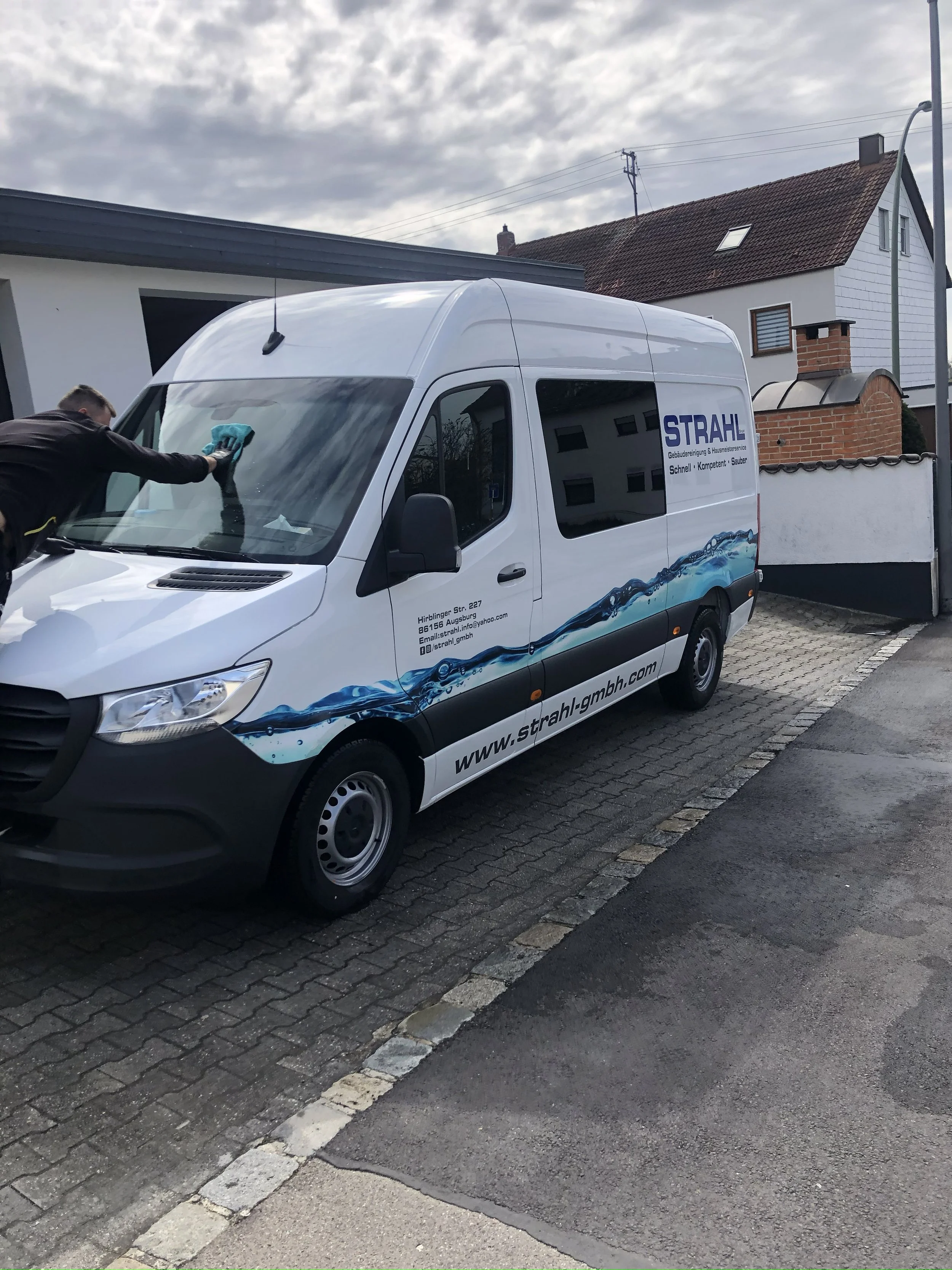 Ein weißer Firmenwagen der Firma STRAHL wird gereinigt, während eine Person mit einem Schwamm den vorderen Bereich des Autos sauber macht. Der Wagen steht auf einem gepflasterten Parkplatz vor einem Haus mit dem Himmel im Hintergrund.