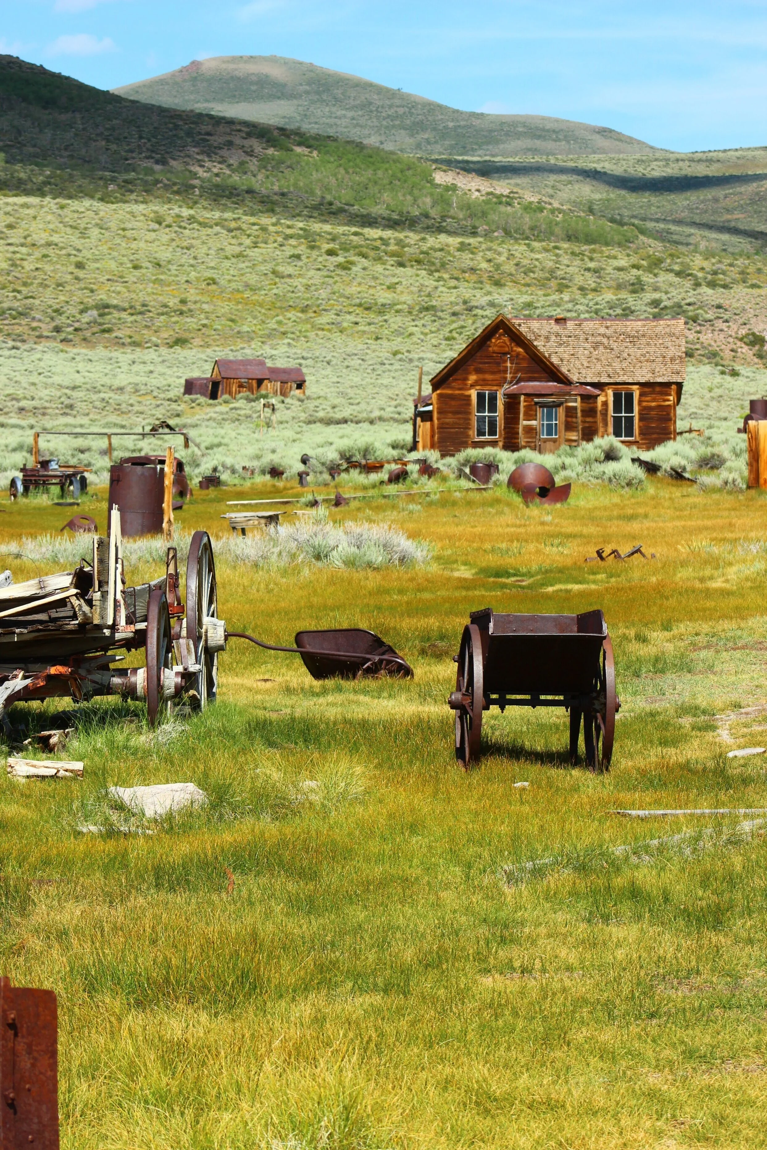 Abandoned farm with old wooden house, rusted farming equipment, and overgrown grass in a rural landscape with hills in the background.
