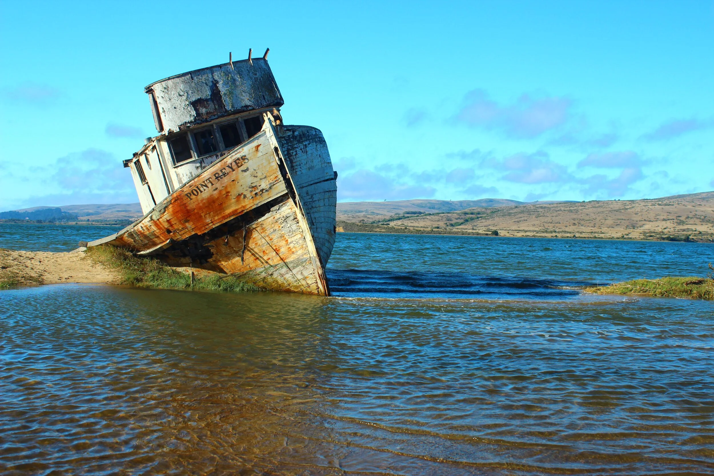 A large, abandoned, rusted shipwreck on the shore of a body of water with hills in the background.