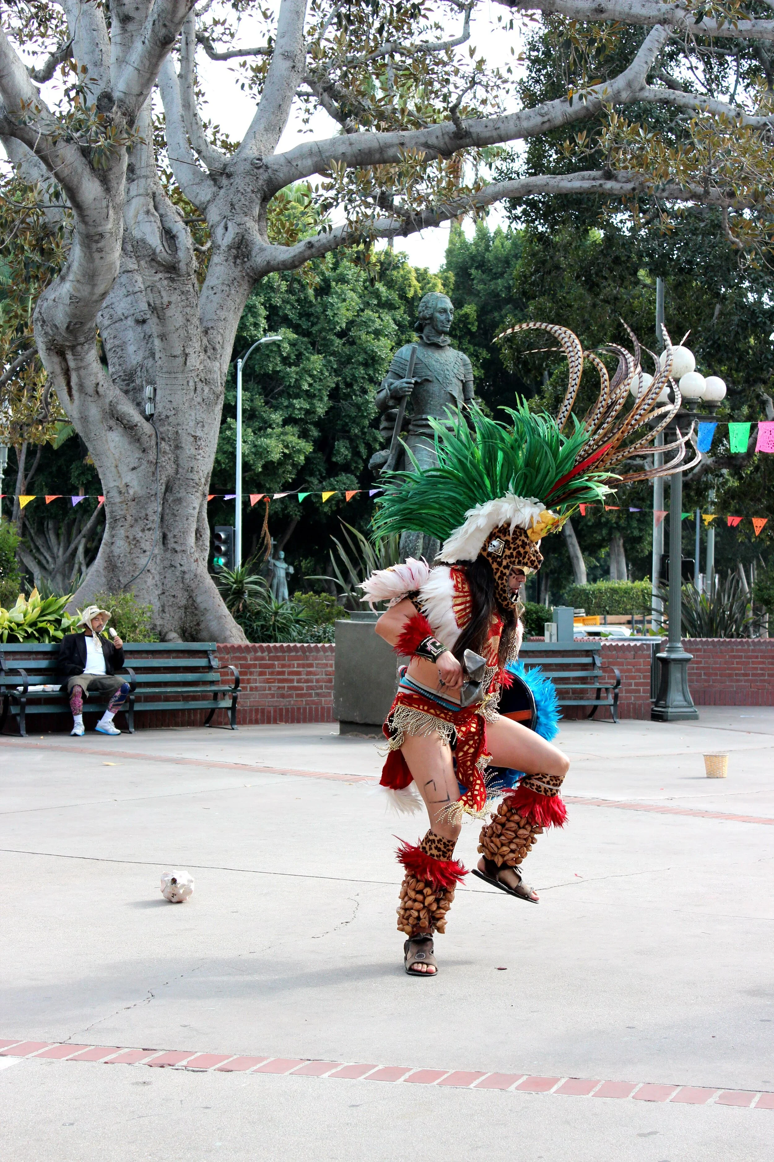 A person dressed in a colorful traditional costume with feathers and animal print is performing a dance in a park, with a large tree, a bust statue, and benches in the background.