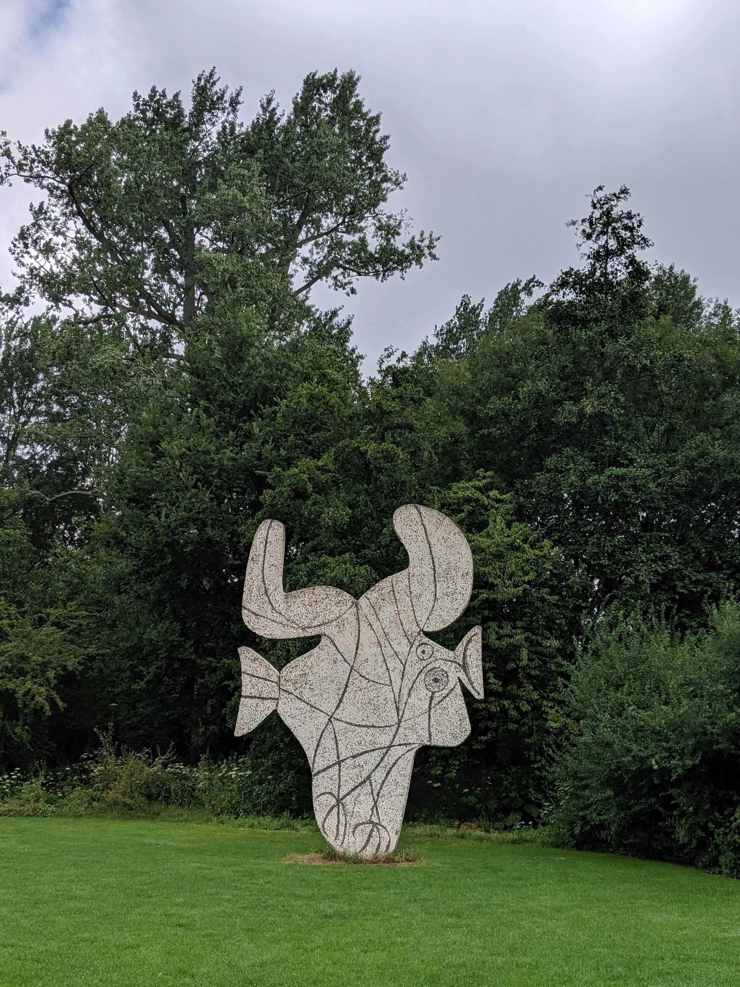 Sculpture depicting a human face with deer antlers and a fish in the background, situated on a grassy area with trees in the background.