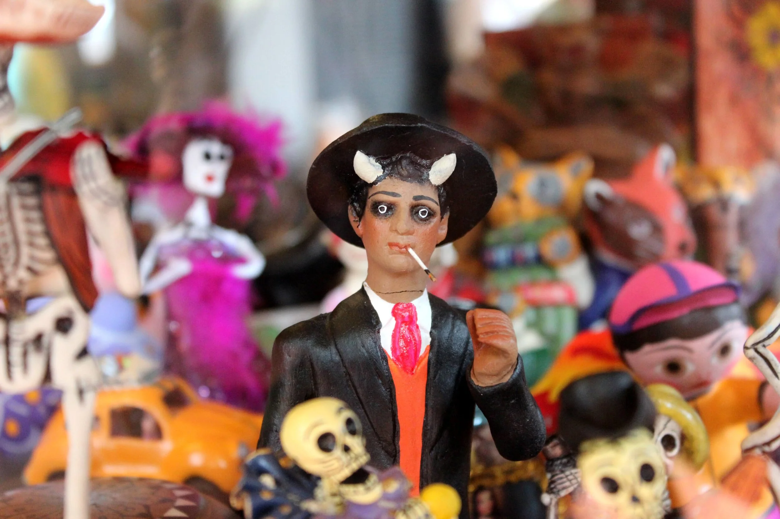 Colorful Mexican Day of the Dead figurine of a person with a cigarette in their mouth, wearing a black hat with white decorations, a black jacket, and holding a skull, surrounded by other Day of the Dead characters.
