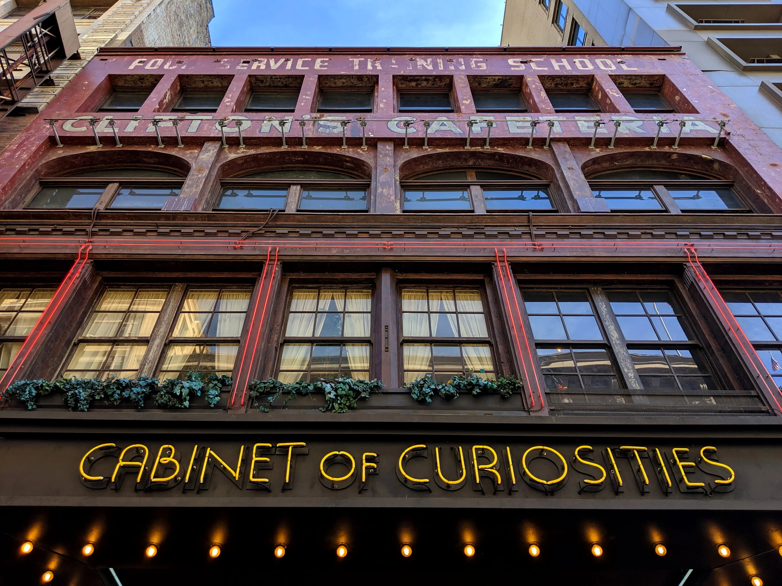 The facade of a multi-story building with vintage signage and neon lights. The top part shows a faded red sign that says 'FOUR SERVICE TRADING SCHOOL' and 'CATERING GALLERY.' The middle features arched windows with greenery below. The bottom has a bl