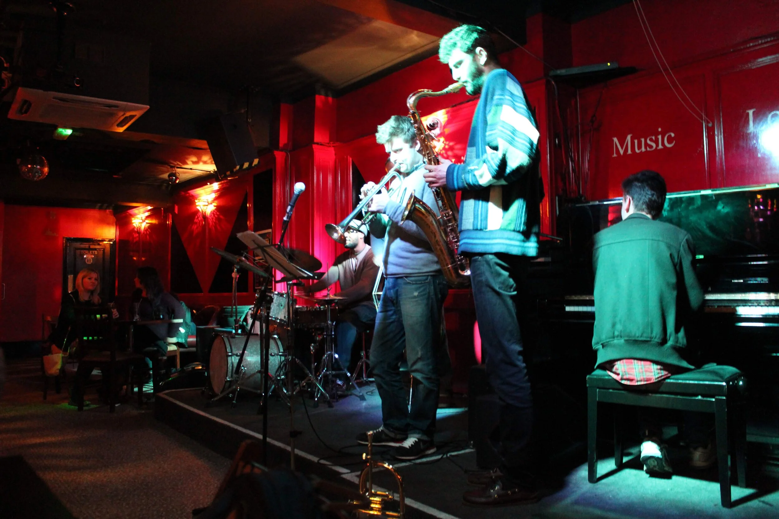 Jazz band performing on stage in a dimly lit red-walled venue, featuring musicians playing drums, trumpet, saxophone, and piano.
