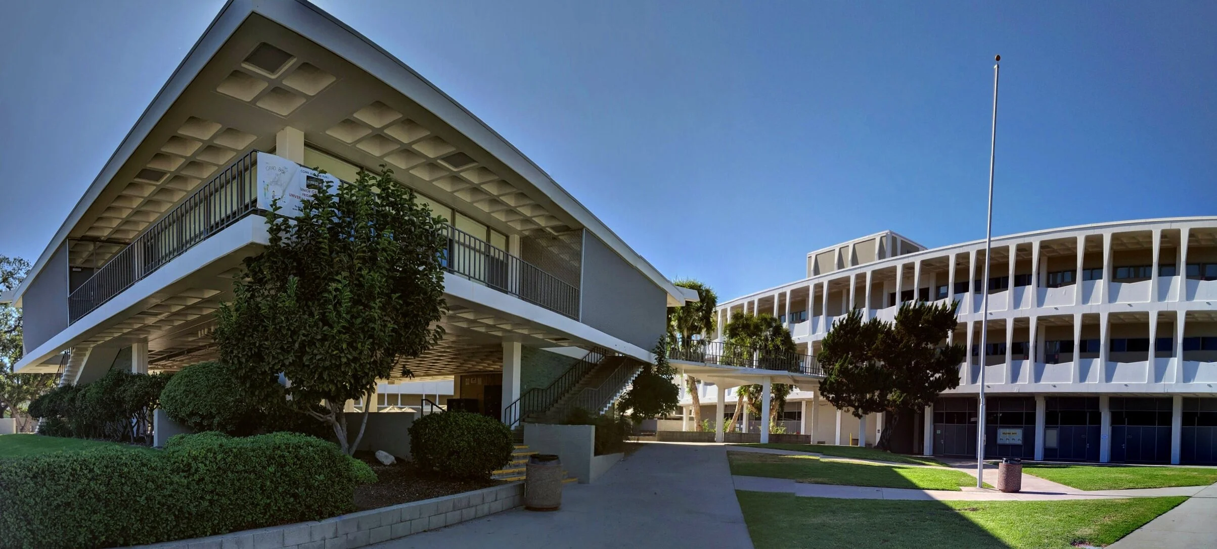 Modern university or office buildings with curved architecture, surrounded by well-maintained lawns, bushes, and trees under a clear blue sky.