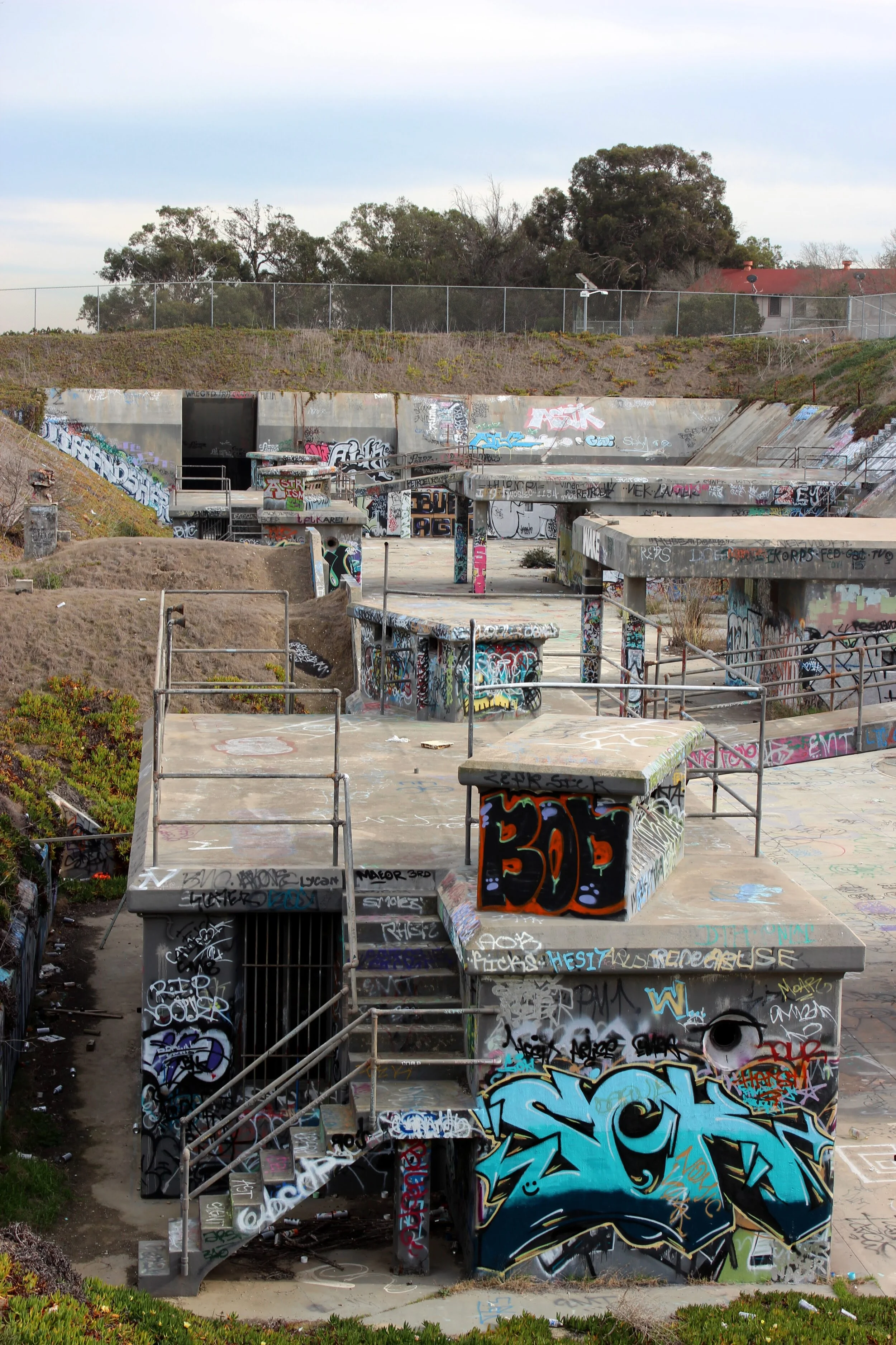 An outdoor skatepark covered in colorful graffiti with various ramps, stairs, and railings, surrounded by greenery and a chain-link fence.