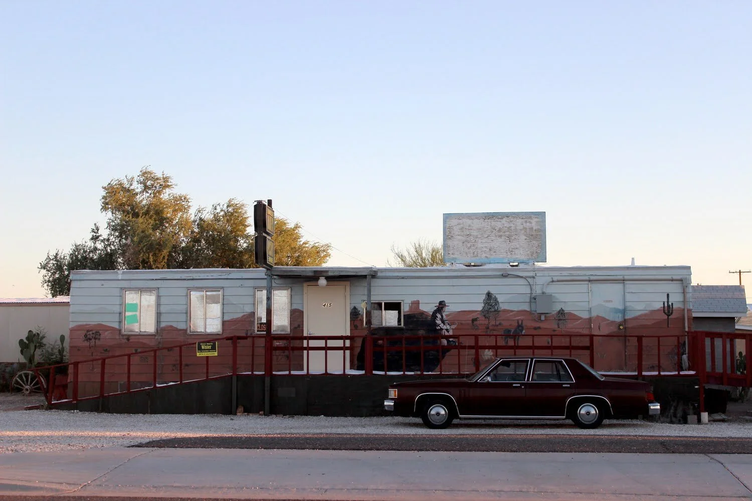 A building with a painted mural depicting a desert landscape, a person riding a horse, and cacti. A maroon vintage car is parked in front, and there is a red railing at the walkway entrance. The sky is clear with a few clouds and trees are visible in