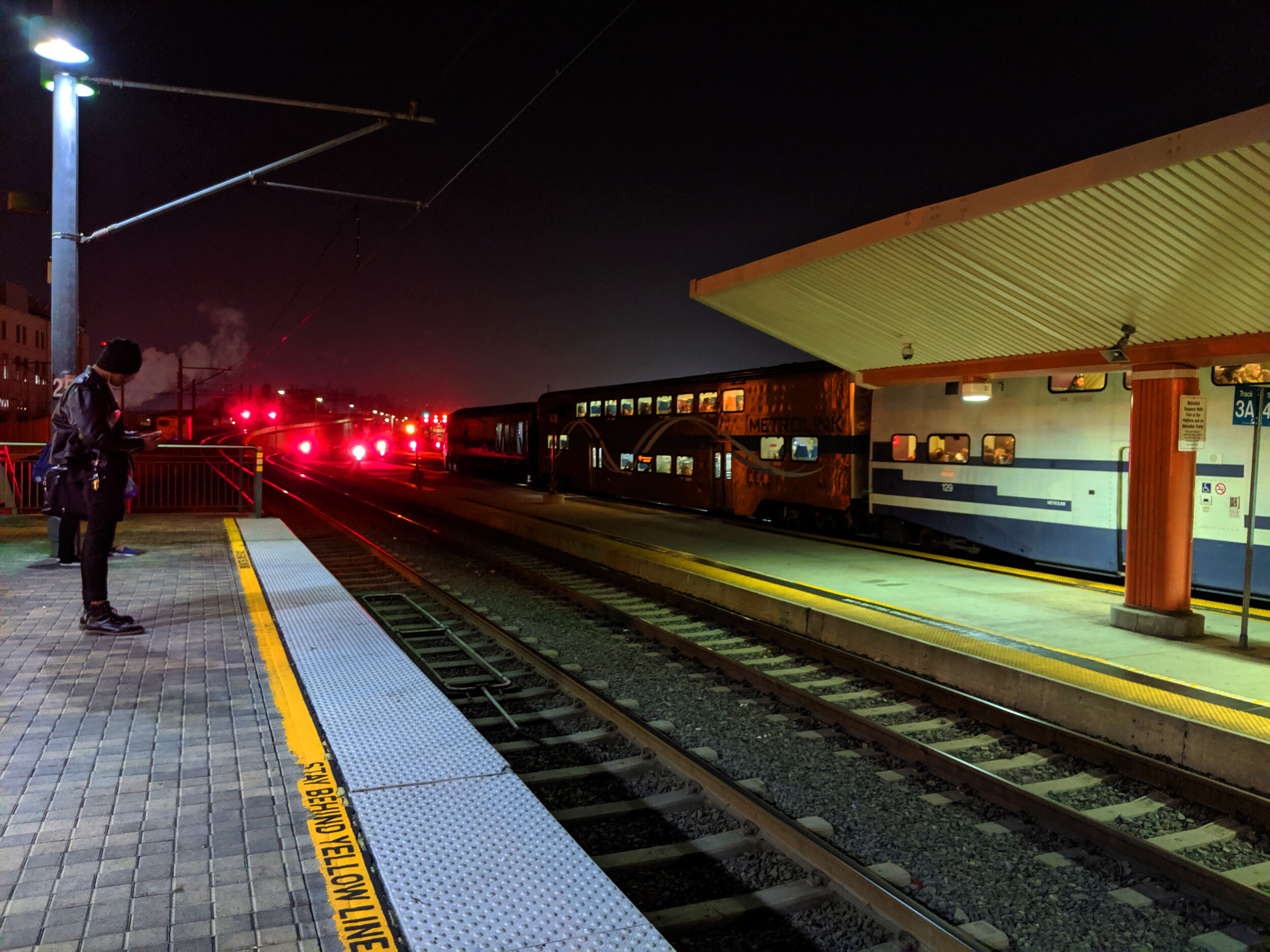 Person waiting on platform at night with train and red signal lights in background.