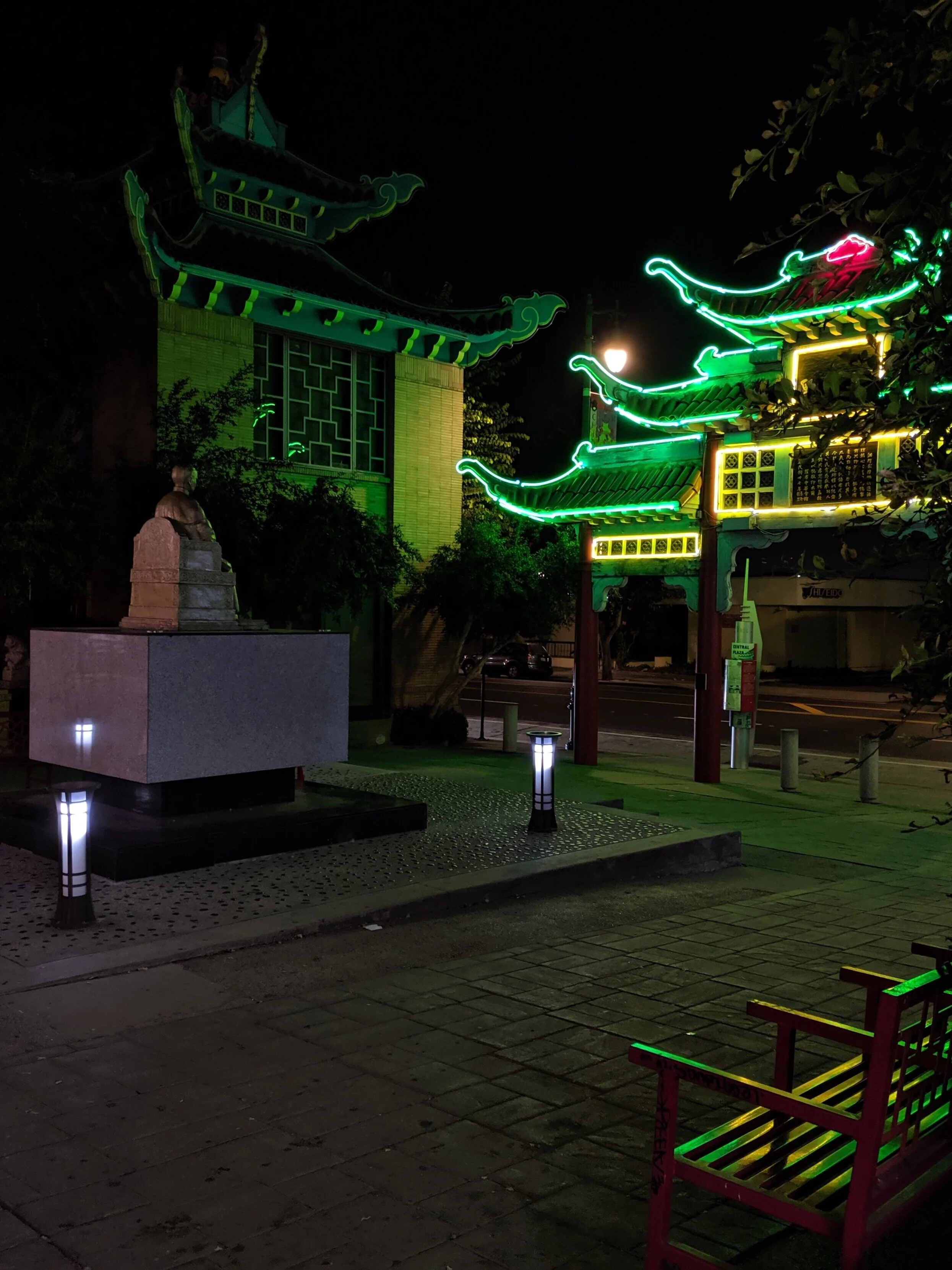 Night scene of a Chinatown gate with neon green and yellow lights, traditional Chinese architectural style, stone statue, small trees, pathway with small lamps, and a red bench.