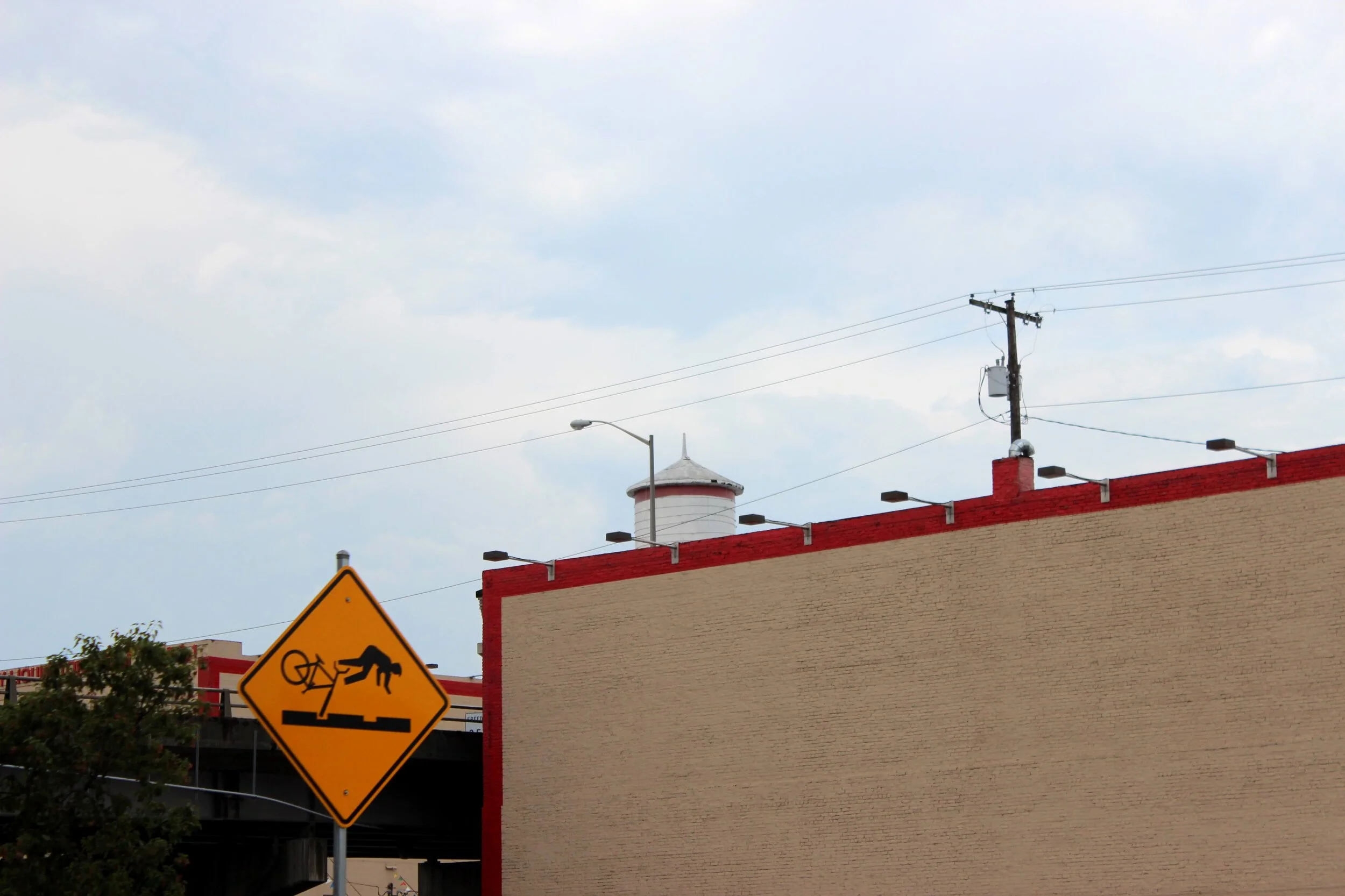 Street scene with a yellow bike accident warning sign, a beige building with a red border, and utility poles with wires against a cloudy sky.
