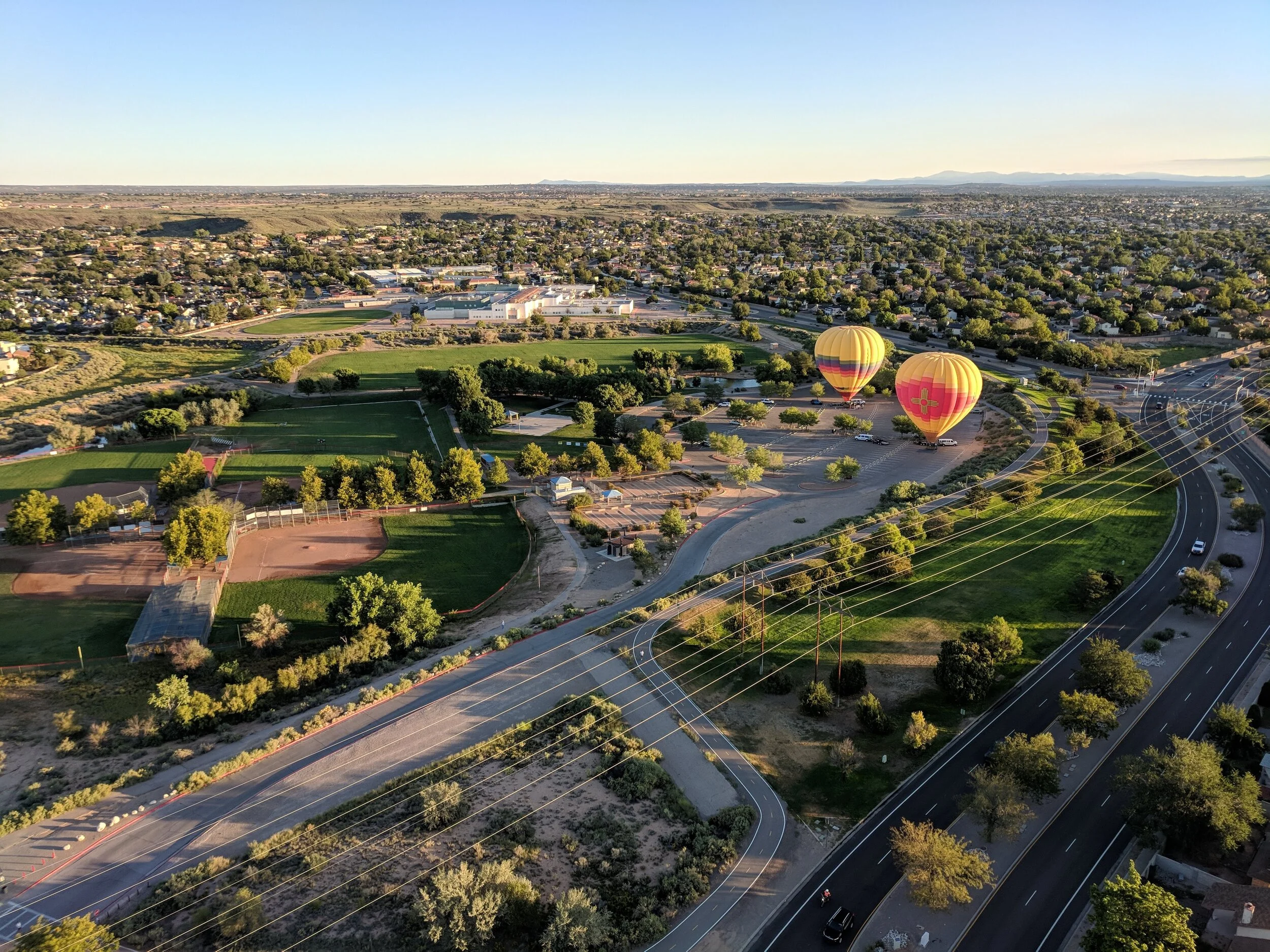 Two yellow hot air balloons with red and pink patterns floating over a park and cityscape at sunset.