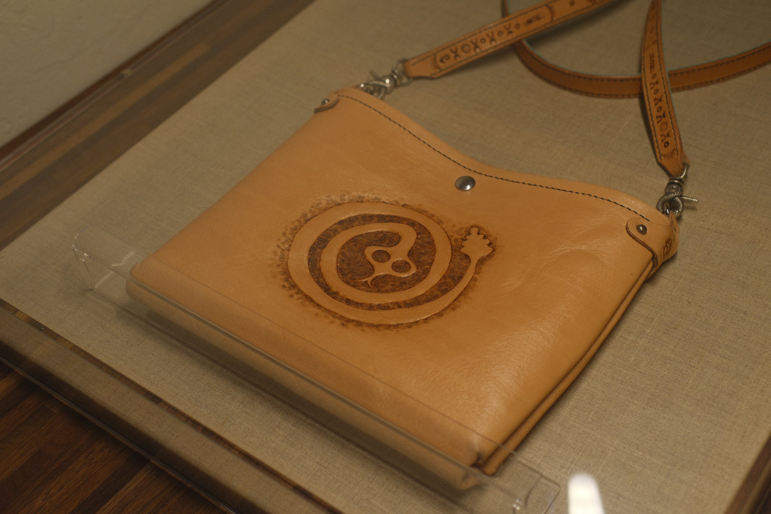 A tan leather shoulder bag with a snake logo and a thin strap displayed in a glass case.