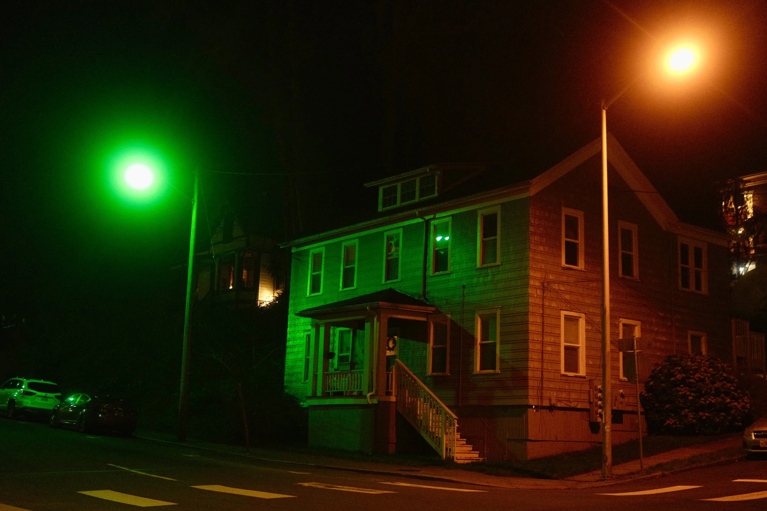 Nighttime scene of a house illuminated with green and orange lights, with streetlights and parked cars visible.