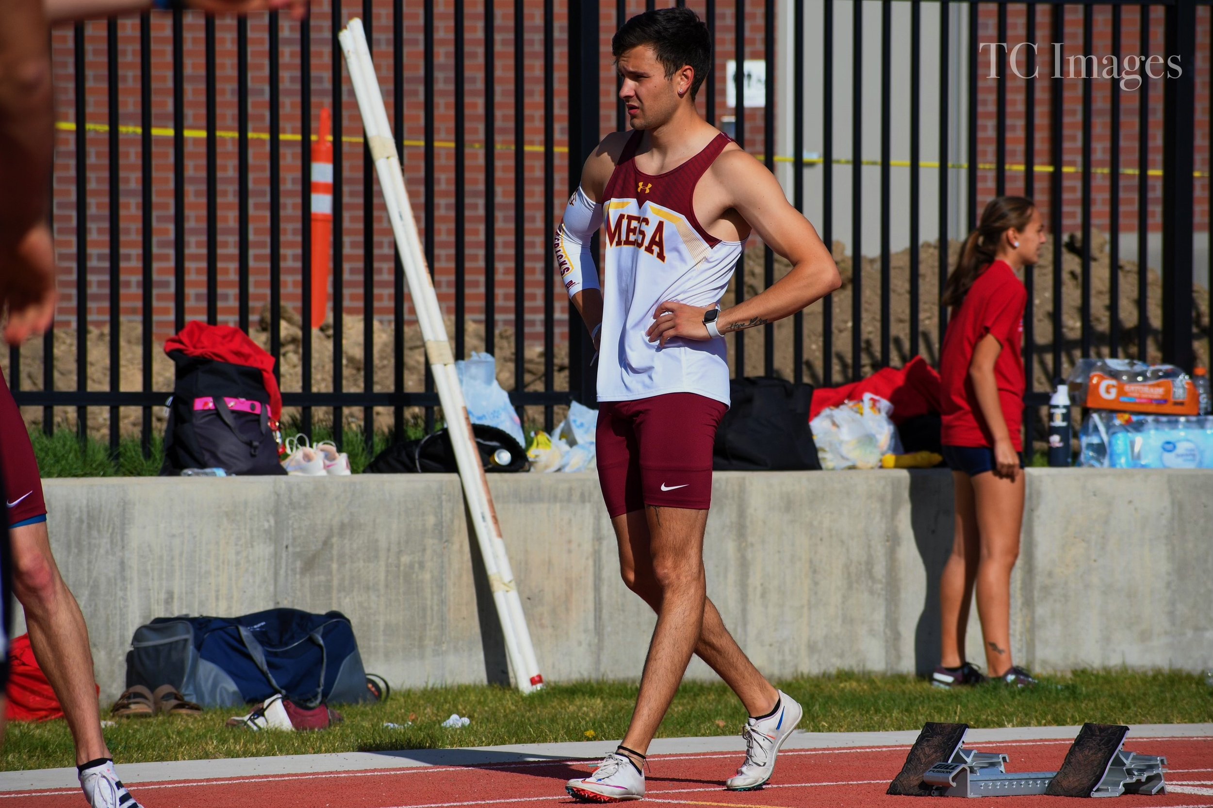 A male track athlete wearing a white and maroon uniform with 'MESA' on the front, standing on a track with his hands on his hips, during a race or practice session.