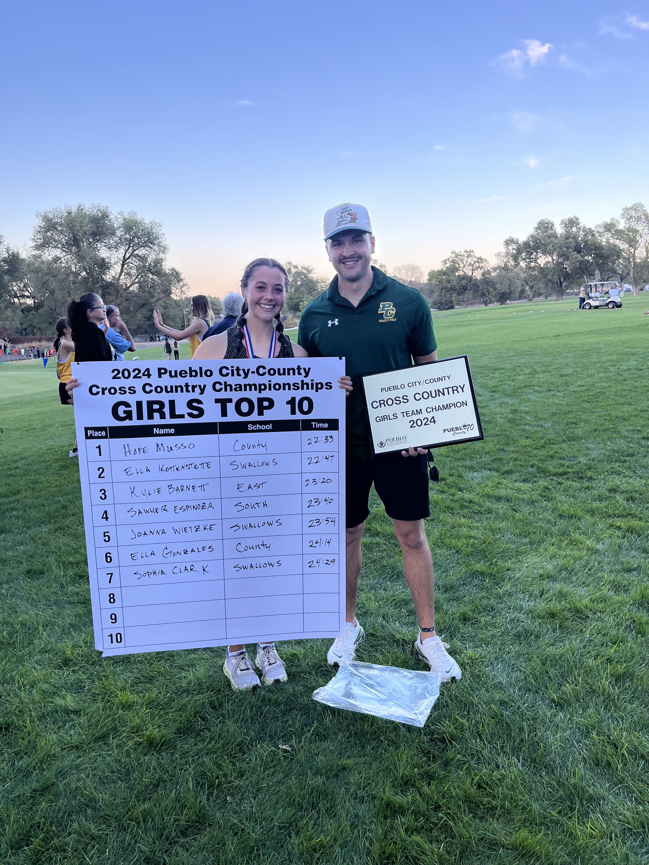 Two people standing on grass holding trophies and a large chart, celebrating at a cross country event with other participants in the background, trees, and a golf cart.