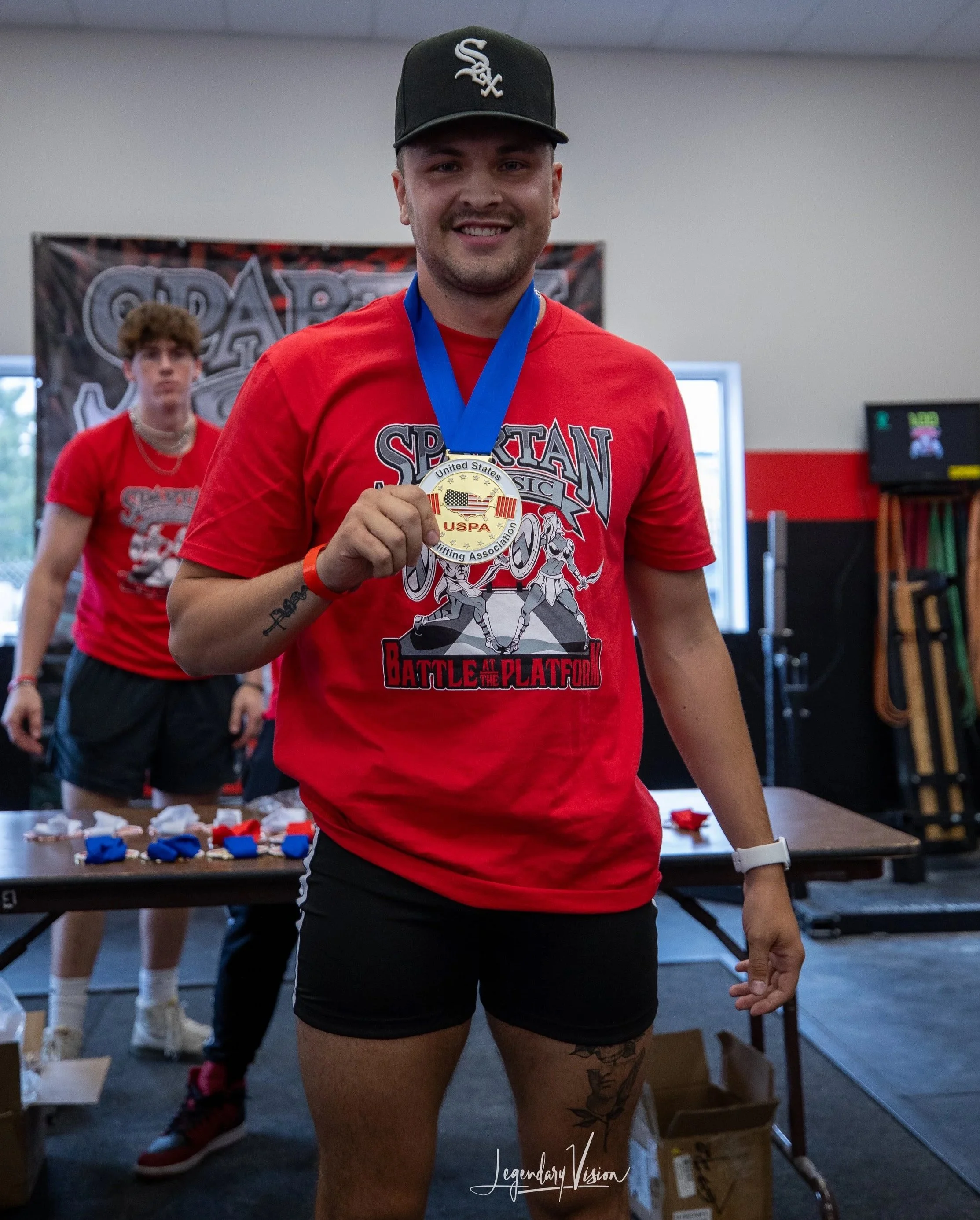 A woman holding a gold medal at a powerlifting competition, wearing a red shirt and black shorts, with another person in the background also in red shirts. The woman has a tattoo on her thigh and is smiling at the camera.