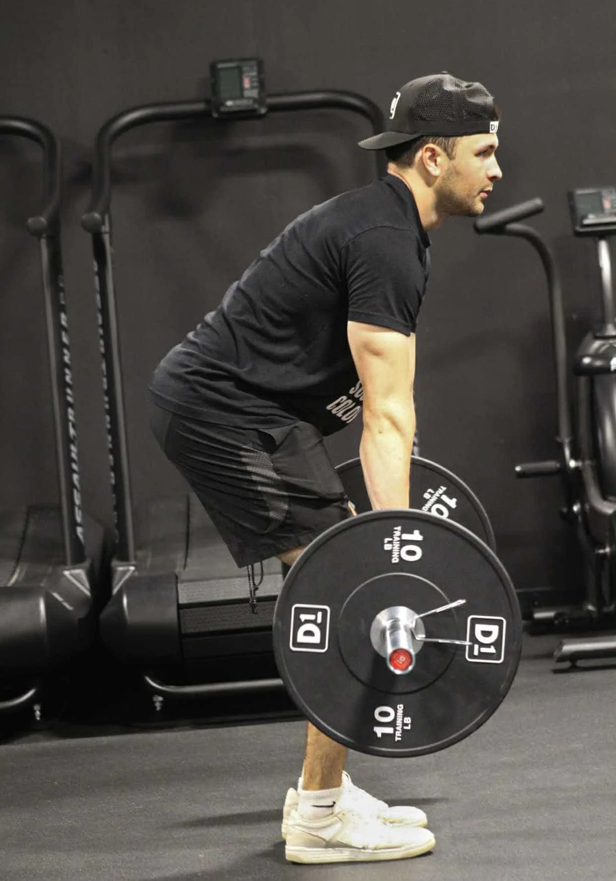 Man in black workout clothes and a black baseball cap performing a deadlift with a barbell in a gym.