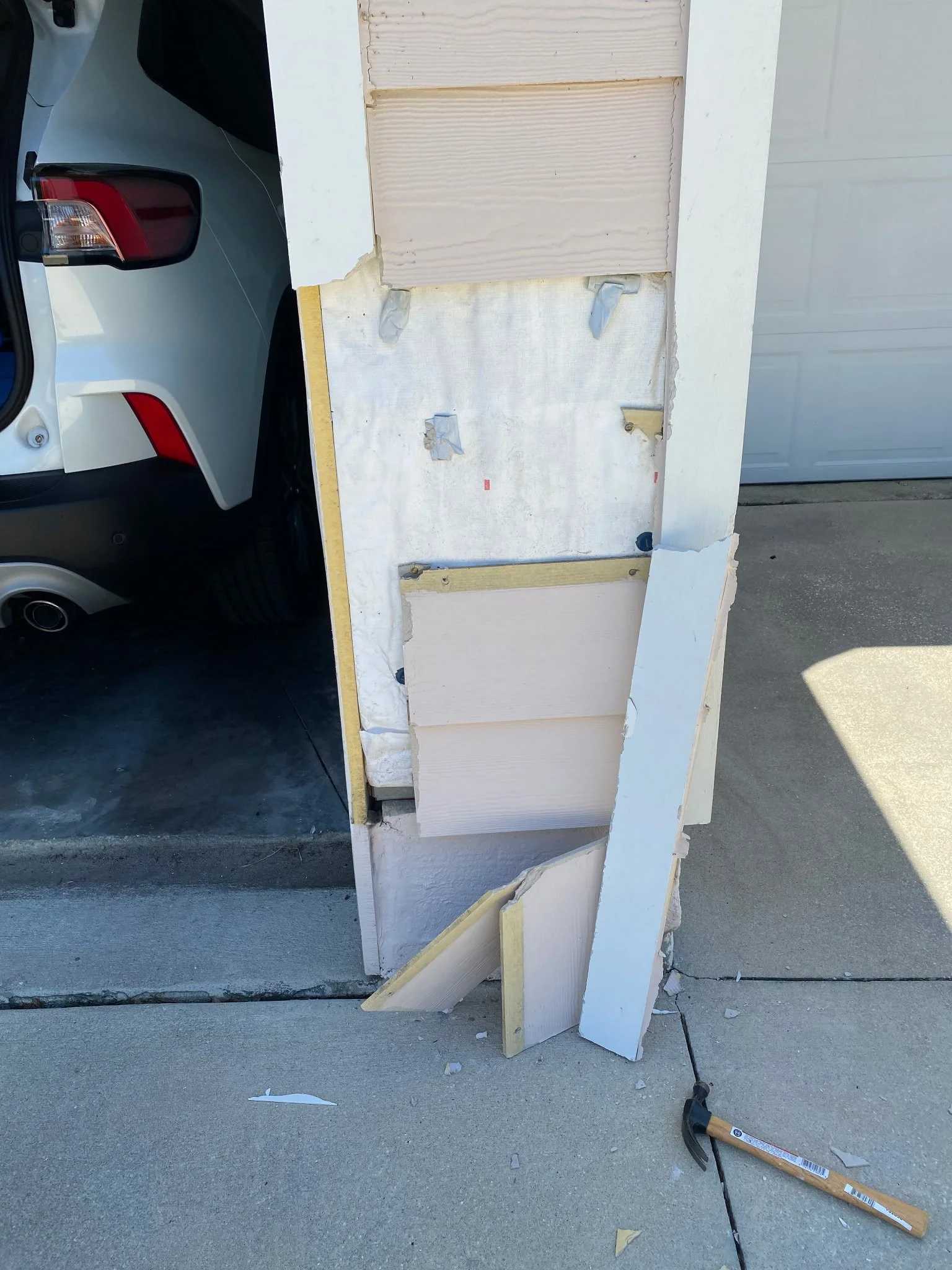 Disassembled siding and insulation panels leaning against a wall, with a hammer on the ground nearby and a car parked in the driveway.
