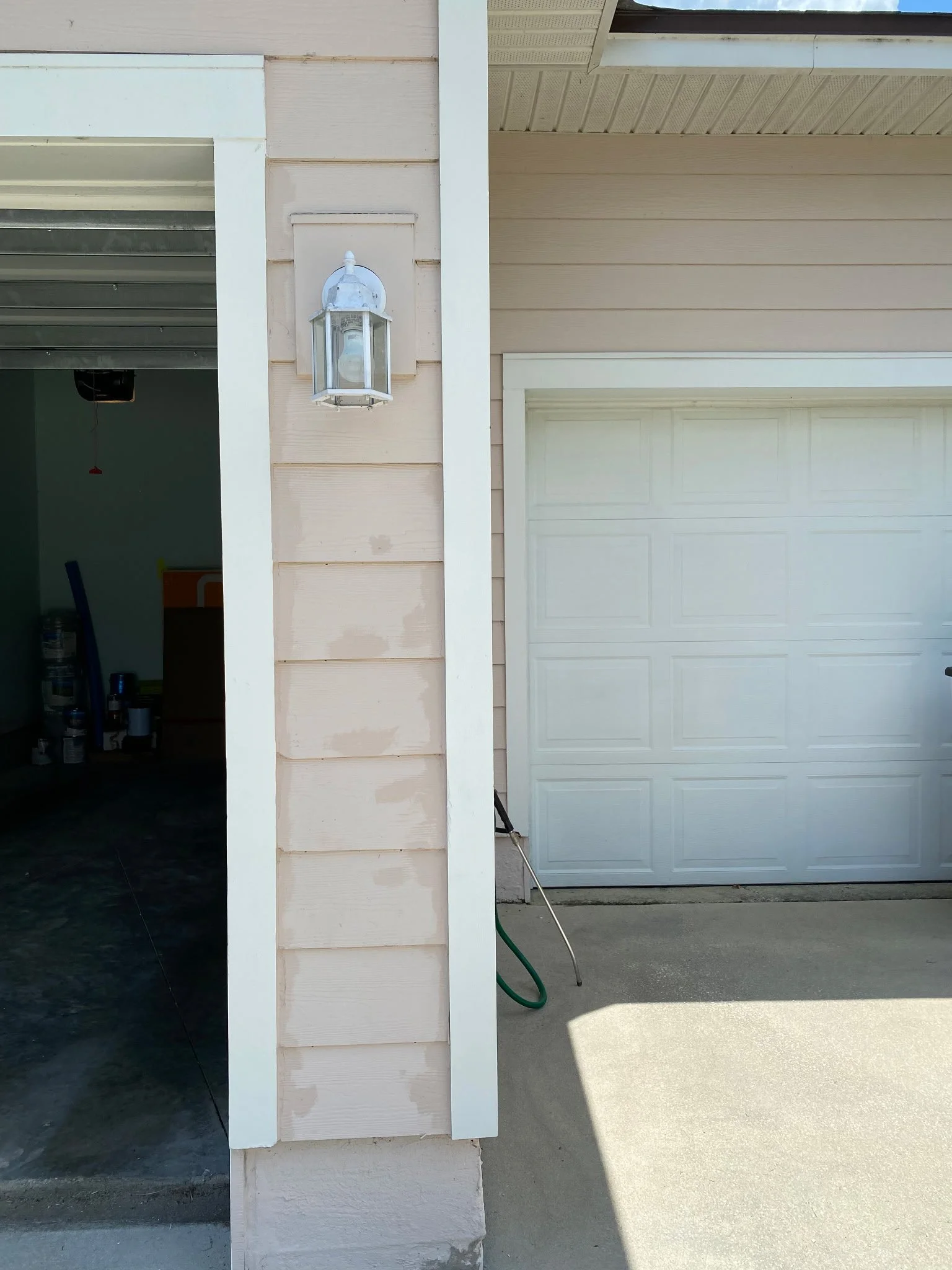 Exterior view of a garage with a white door, beige siding, and a lantern-style light fixture on the wall. Some garage supplies visible inside.