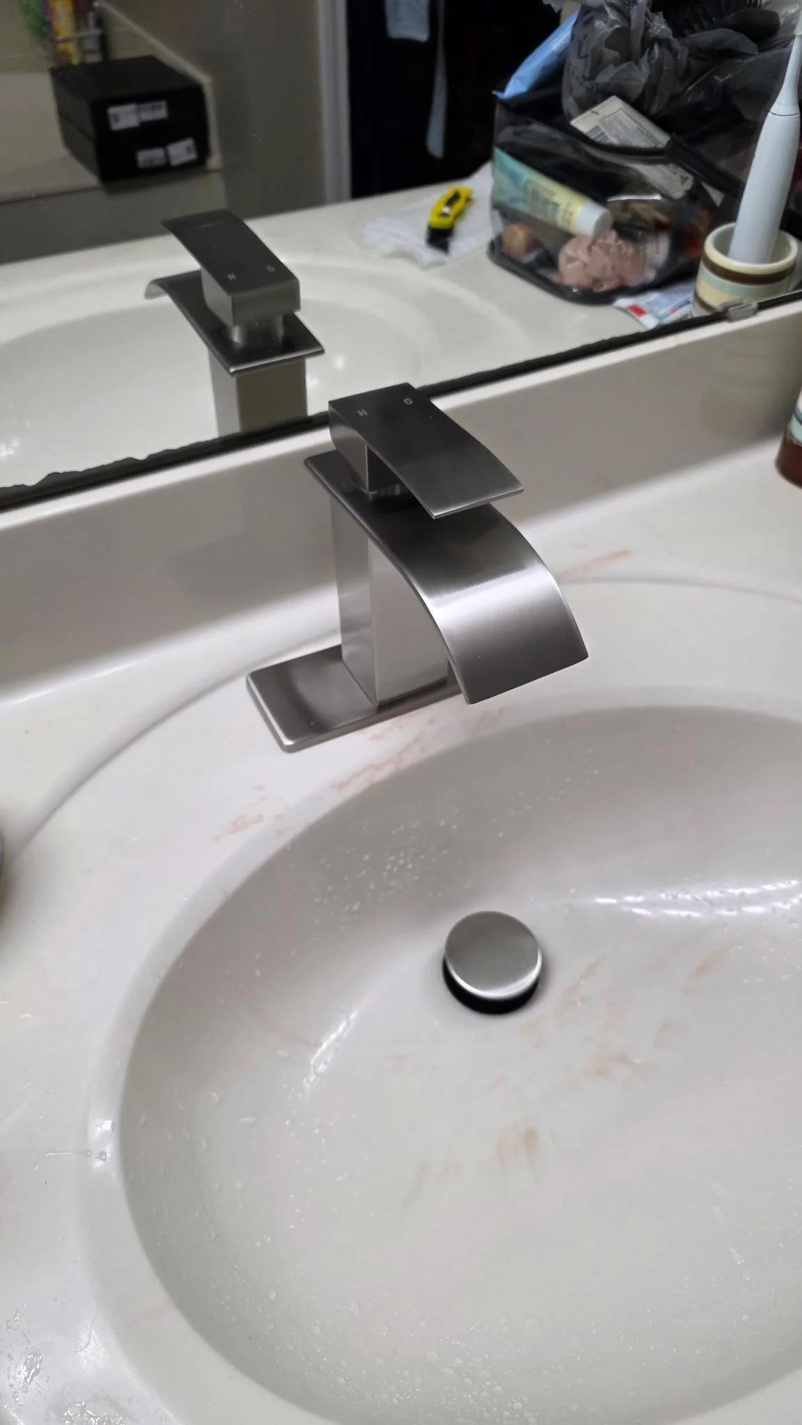 Modern silver bathroom faucet above a white sink with soap residue, with a mirror reflecting various toiletries, a container with a cloth, and a yellow utility knife in the background.
