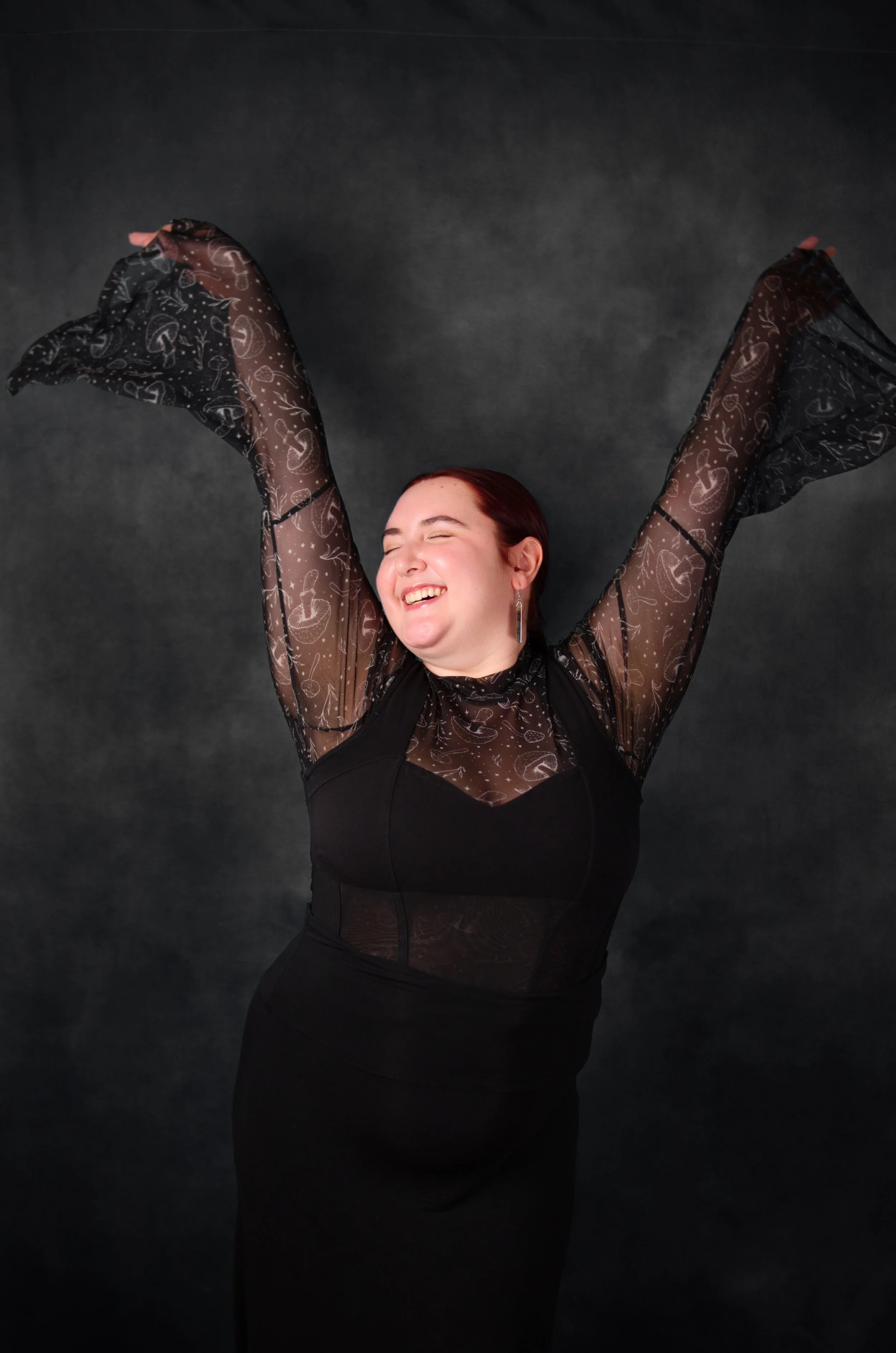 A woman with red hair smiling and stretching with arms raised, wearing a black dress with sheer, patterned long sleeves against a dark background.