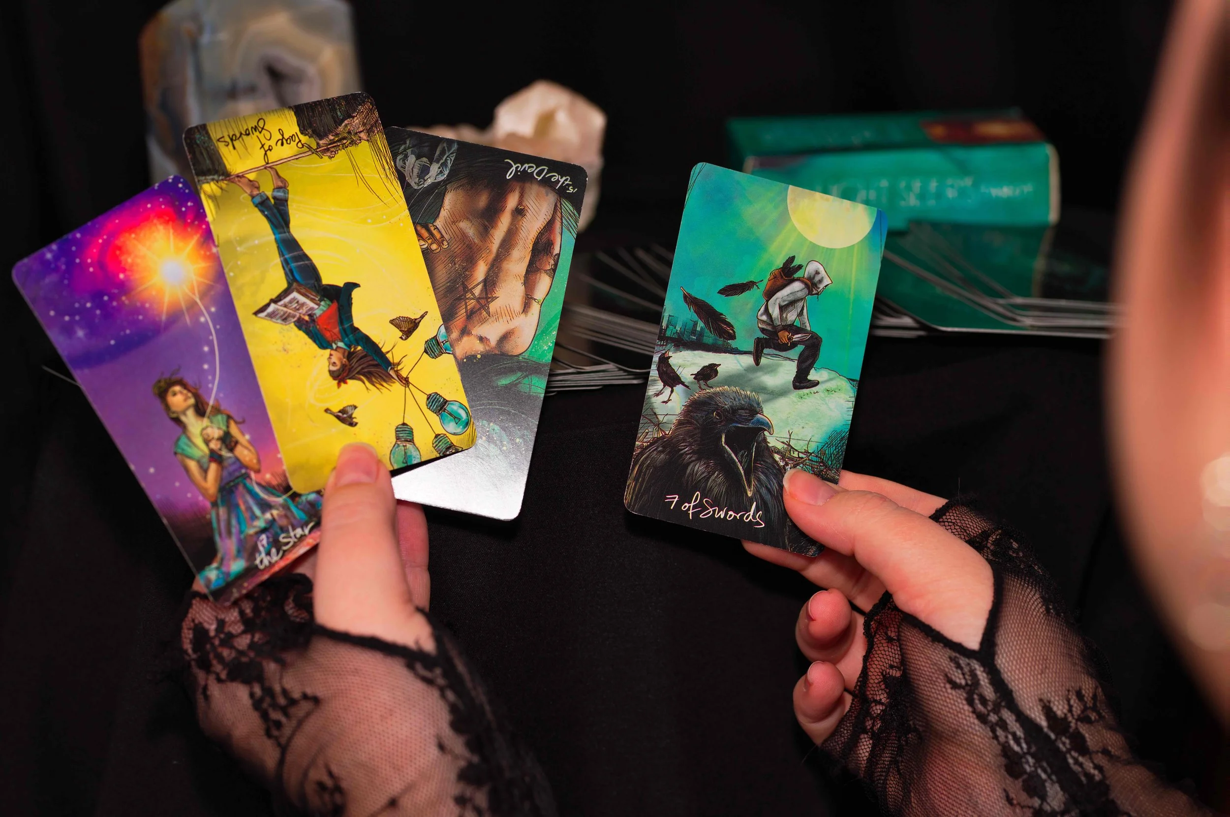 Person holding tarot cards, including 'The Star' and 'Seven of Swords', with a tarot deck in the background, on a black surface, wearing lace gloves.