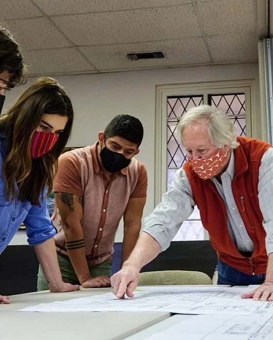 A group of people, all wearing face masks, gathered around a table looking at large sheets of paper with diagrams or plans, indoors with a window and framed picture on the wall.