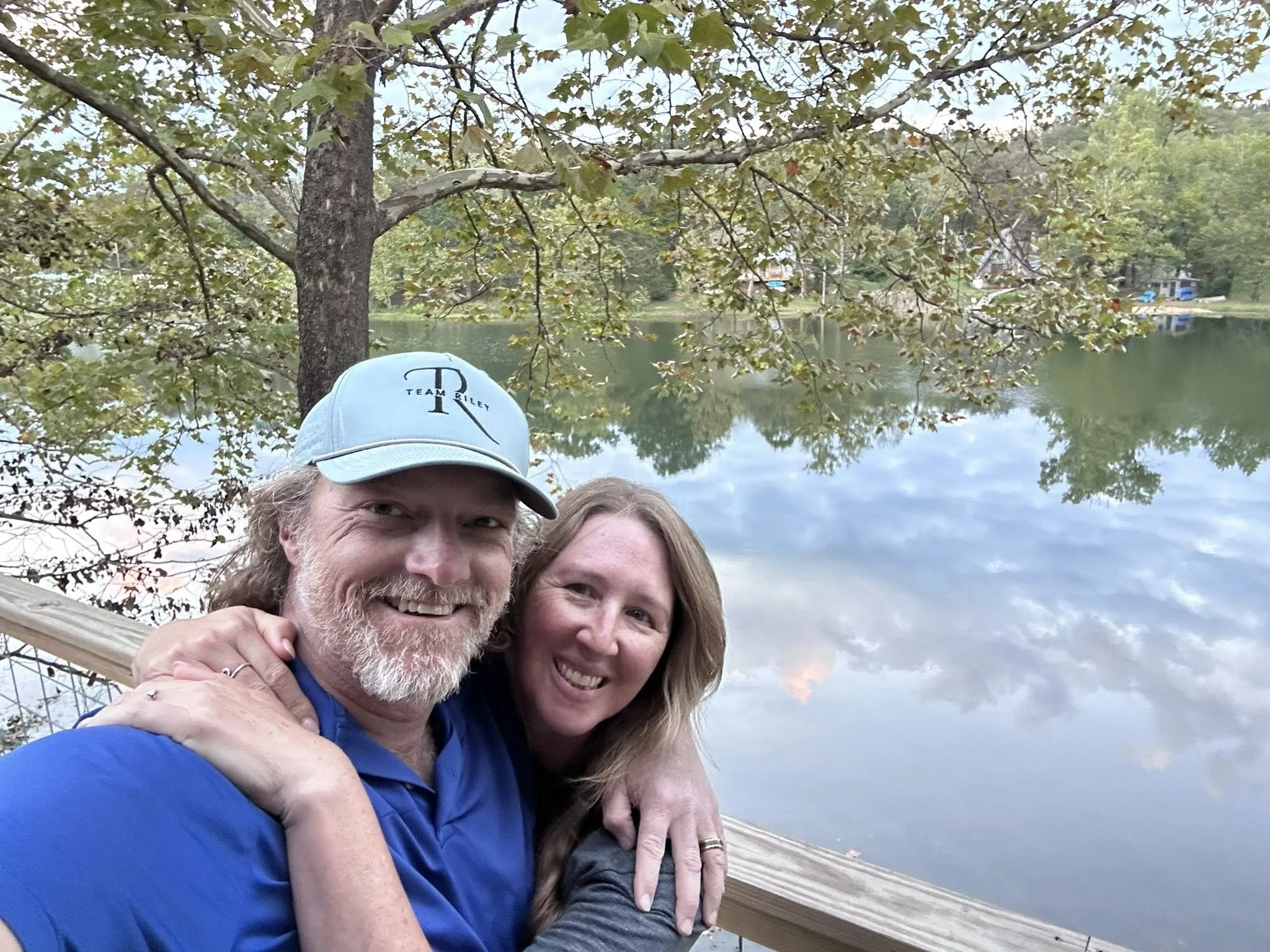 A smiling couple taking a selfie outdoors by a lake during daytime, with trees and clouds reflected in the water.