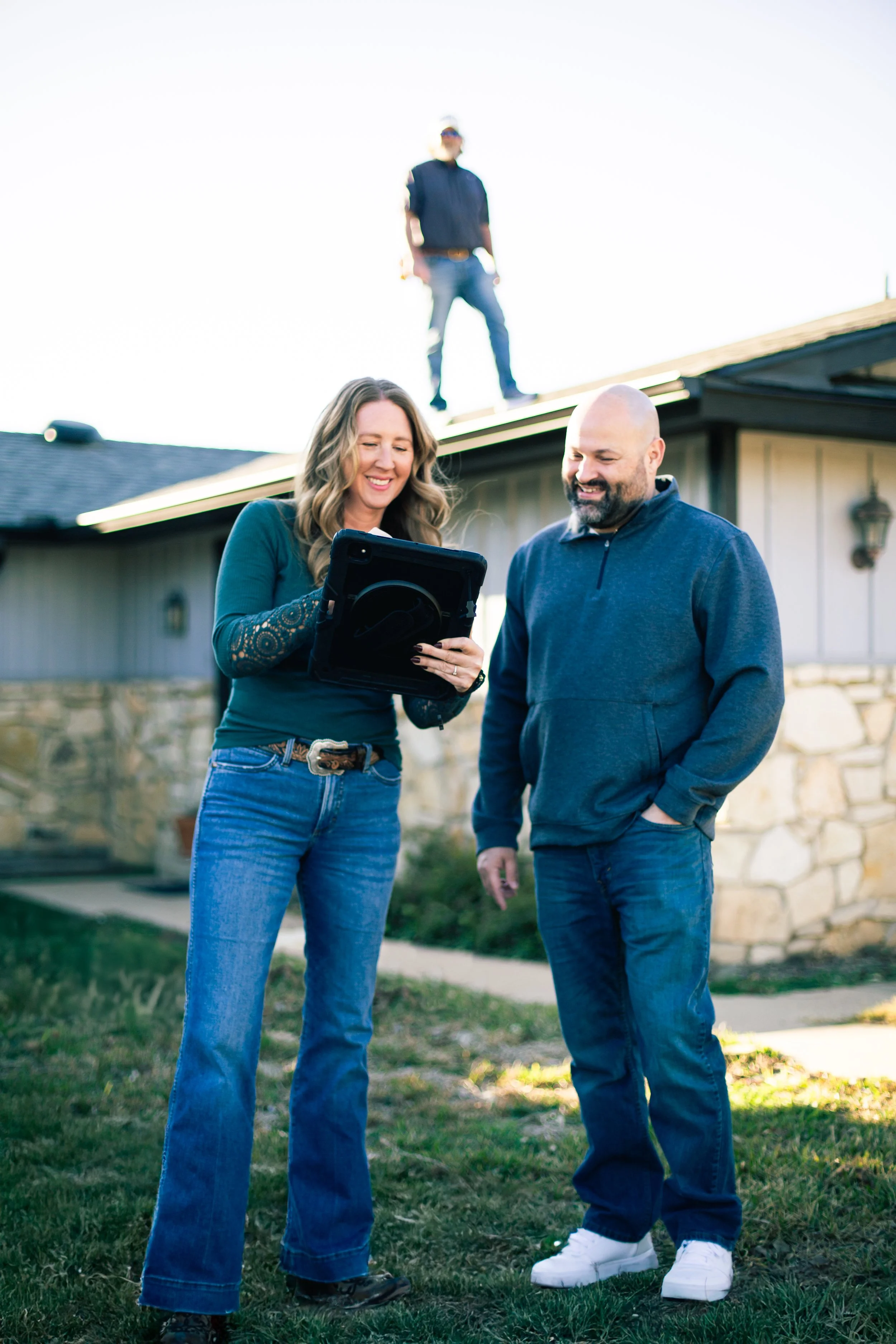 Two people, a woman with wavy hair in a green shirt and jeans, and a man with a beard in a blue jacket and jeans, standing on grass in front of a house. They are looking at a tablet, smiling, while a person in a black vest and jeans stands on the roof of the house in the background.