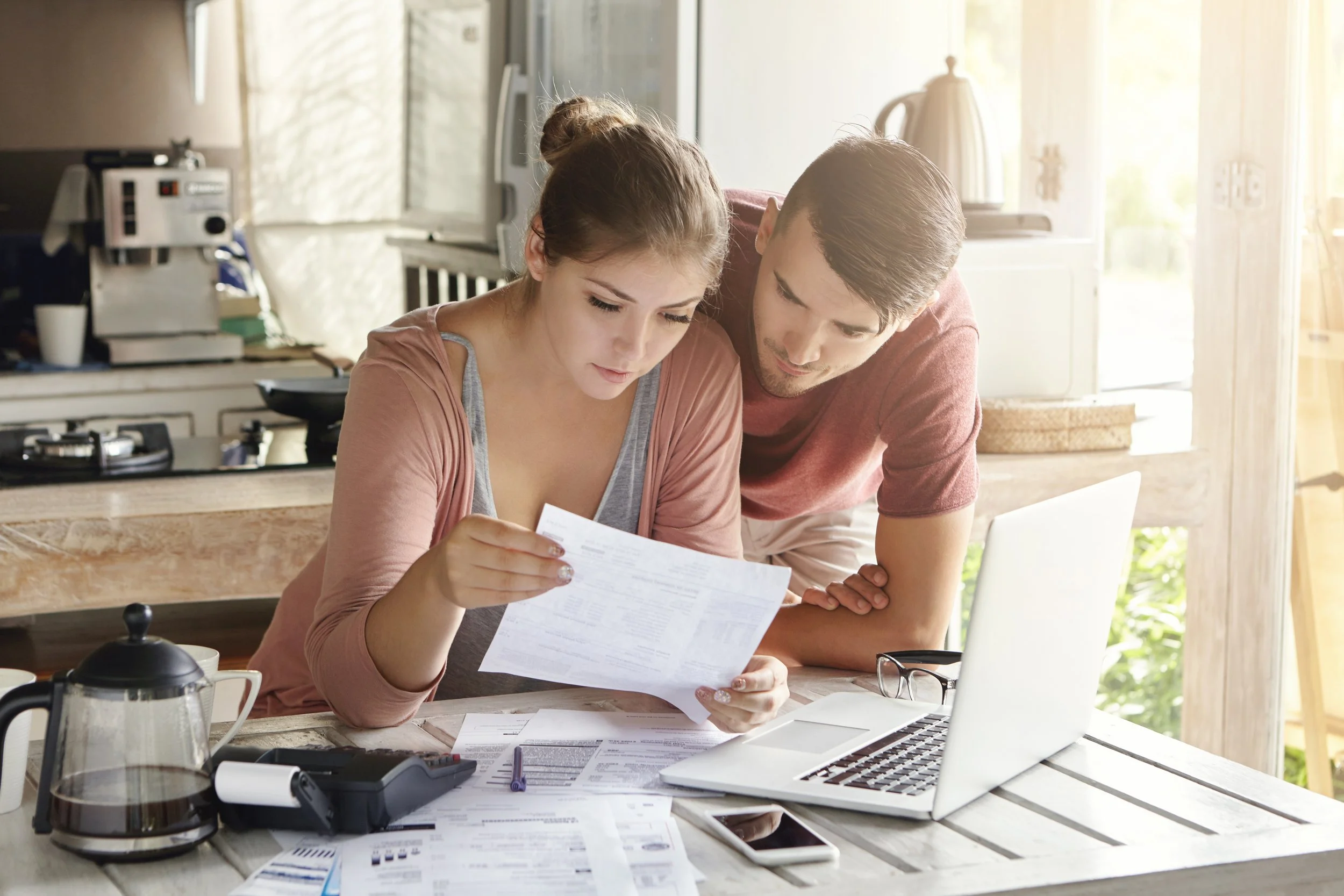A young couple looking at documents on a kitchen table with a laptop, coffee pot, and calculator on the table, sunlight coming through the window.