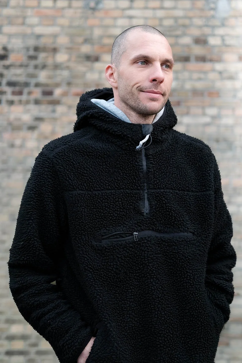 Ein Mann mit kurzen Haaren trägt einen schwarzen, flauschigen Pullover mit Reißverschluss vor einer Ziegelmauer.