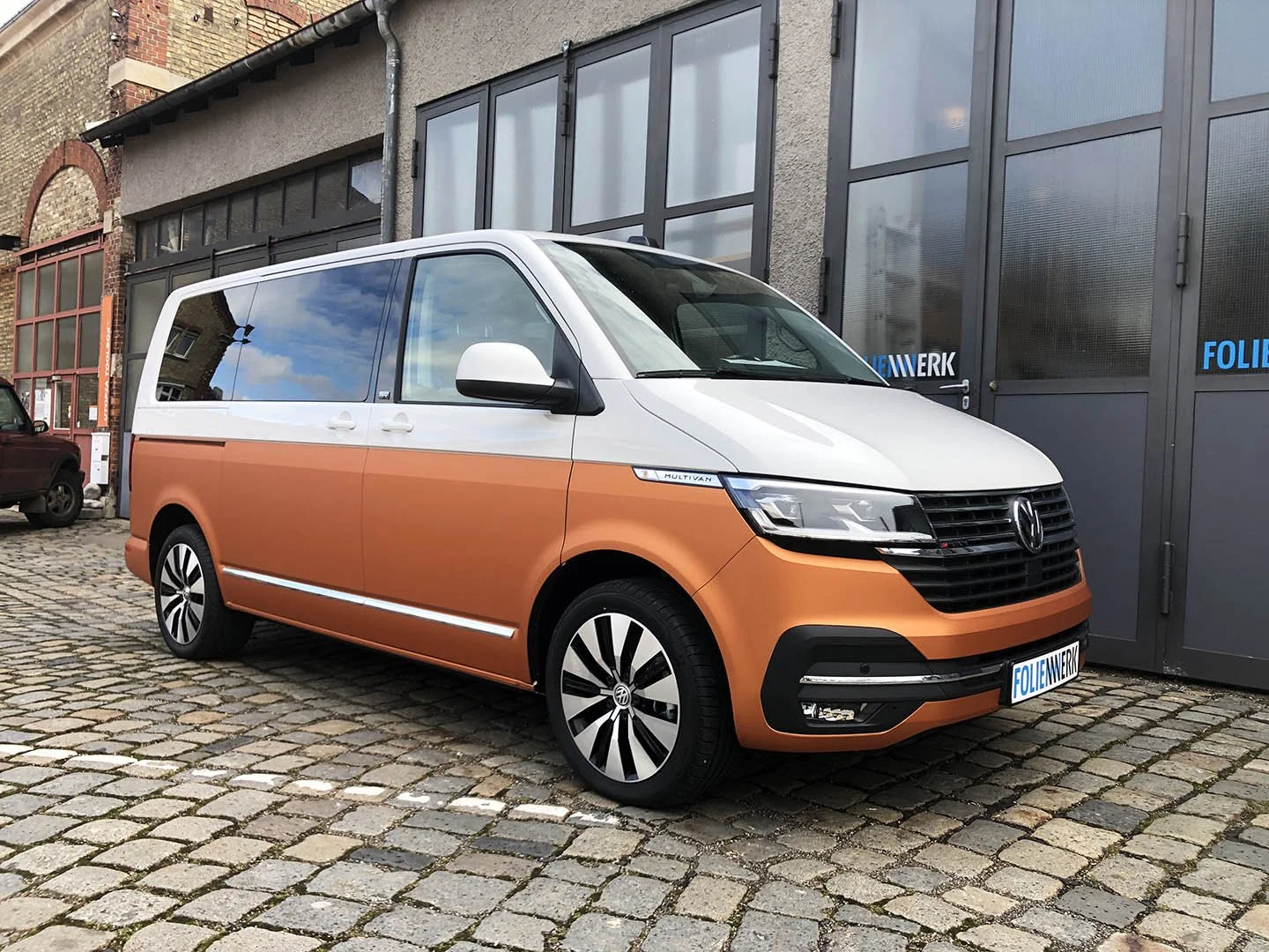 Ein Volkswagen Multivan mit orangenem unteren Bereich und weißem oberen Bereich, geparkt auf einem gepflasterten Platz vor einem grauen Gebäude.