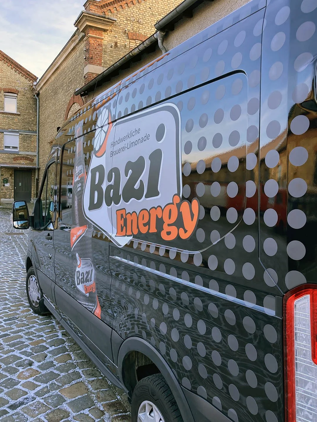 Werbetruck für Bazl Energy mit Logo, orange und grauen Punkten, in einer Straße aus Kopfsteinpflaster, alte Gebäude im Hintergrund.